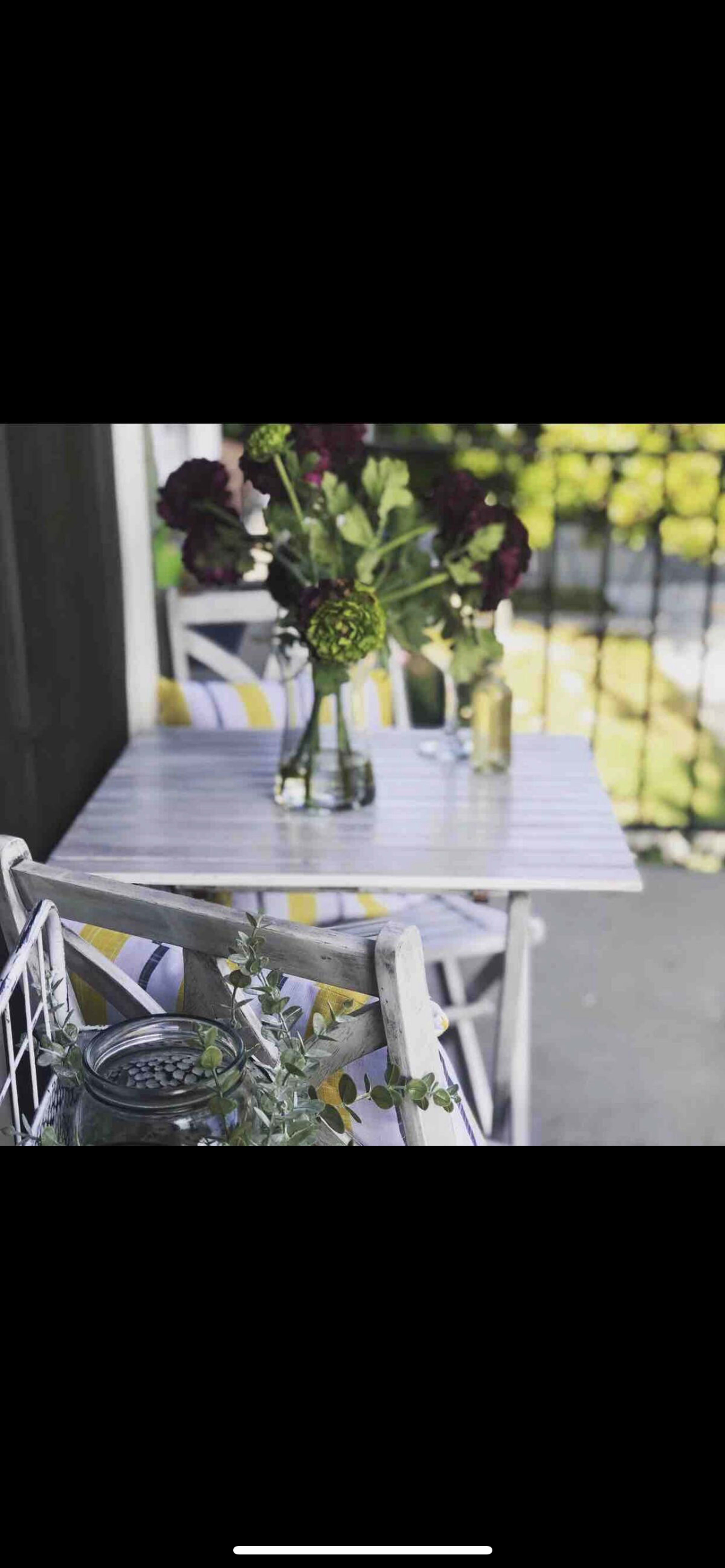 A small, white-striped table is set on a porch, adorned with a vase of flowers and accompanied by colorful chairs and a glass jar.