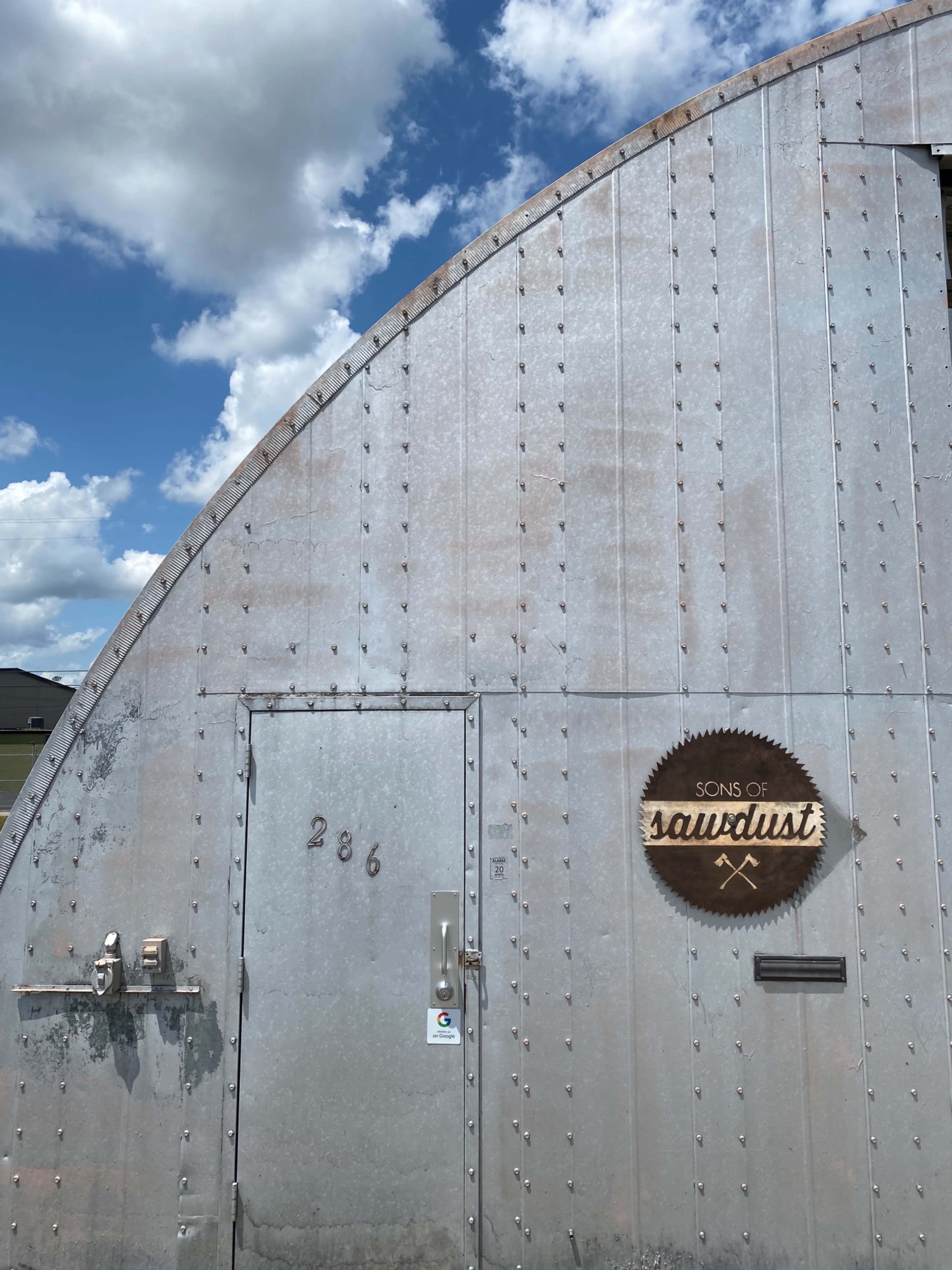 A corrugated metal building features a wooden sign reading "Sons of Sawdust" near a metal door.