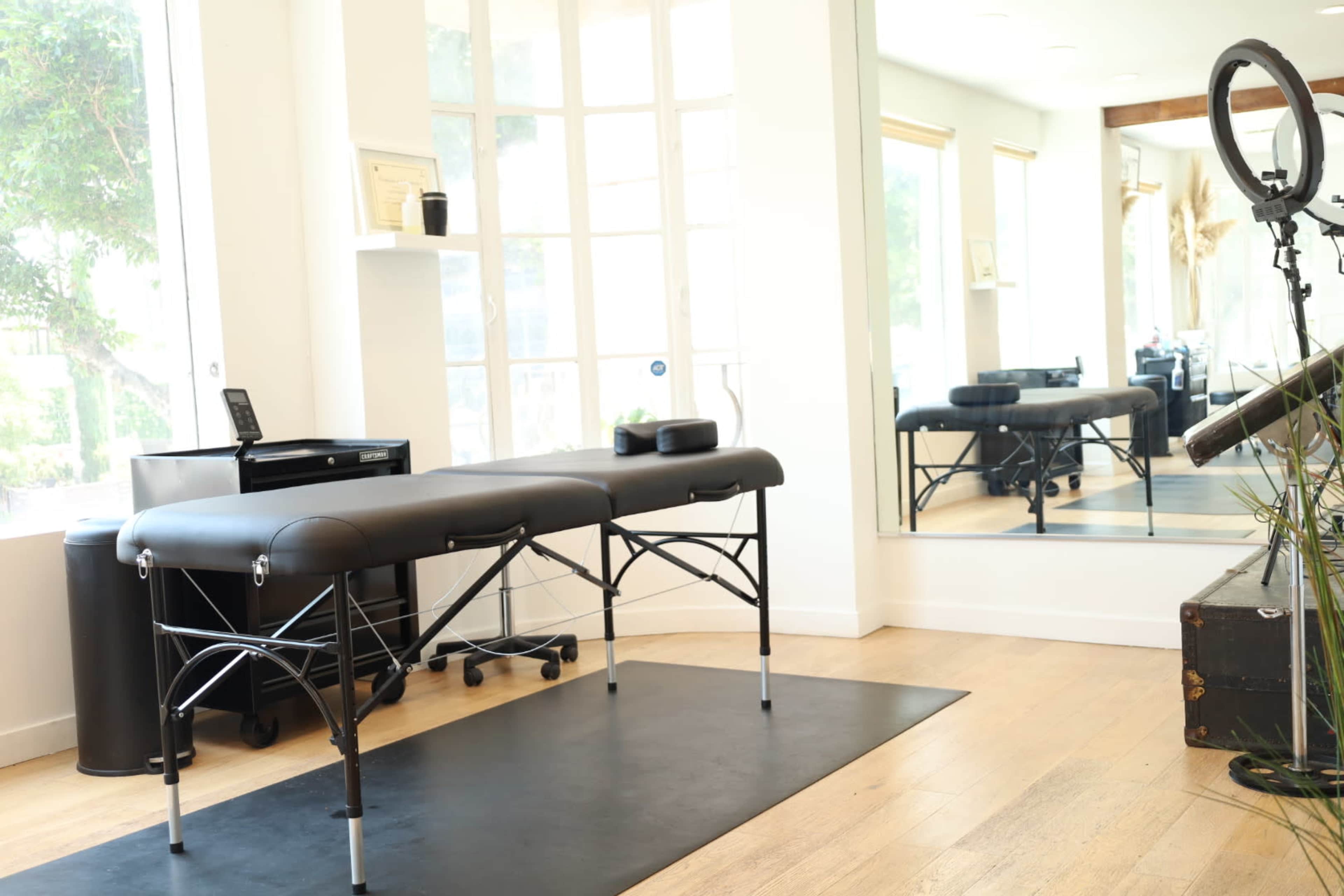 The image shows a massage table set up in a bright room with large windows and a mirror reflecting the space.