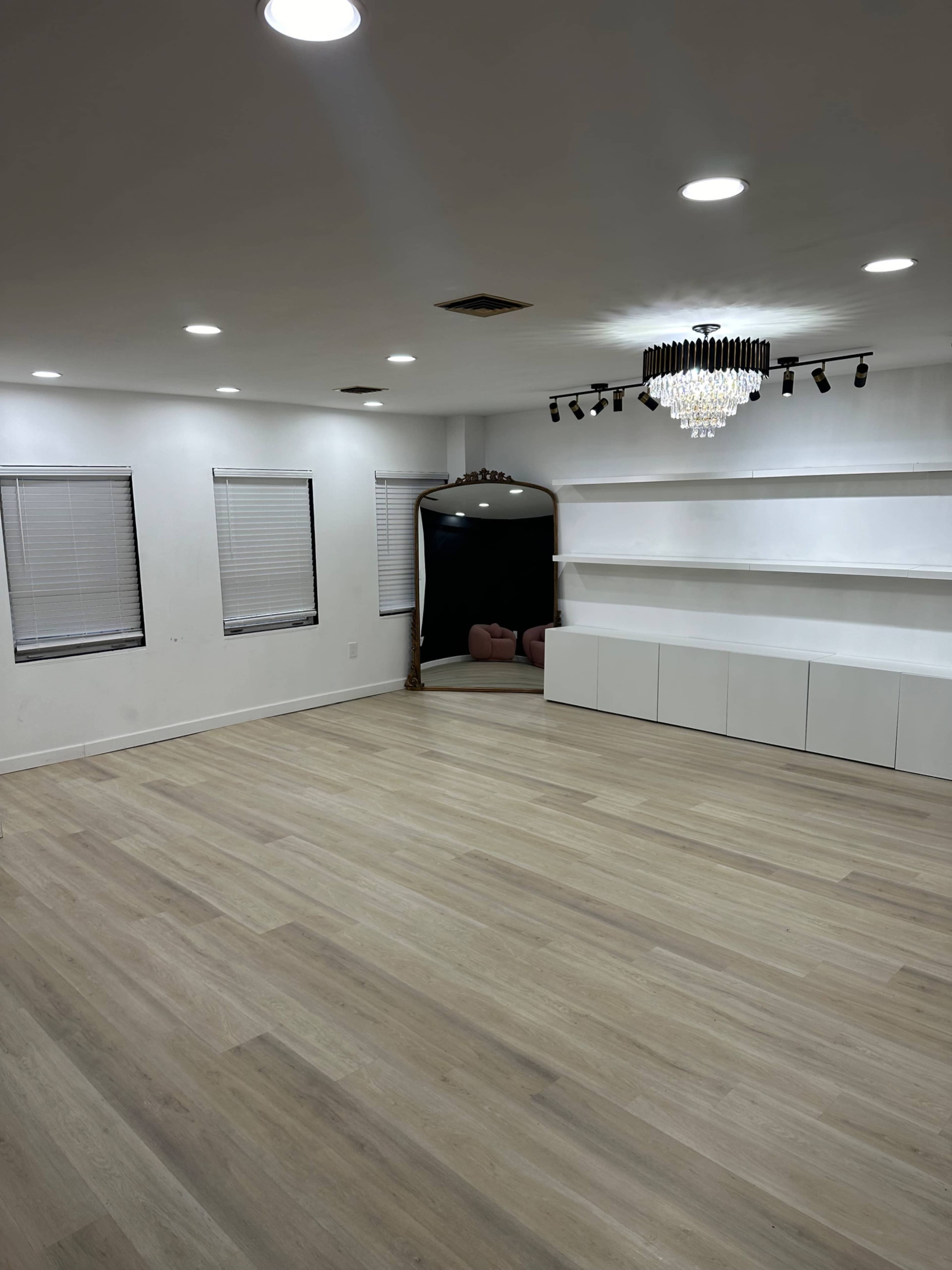 The image shows a spacious room with light wooden flooring, white walls, a chandelier, and shelves along one wall, with a dark mirror reflecting a section of the room.