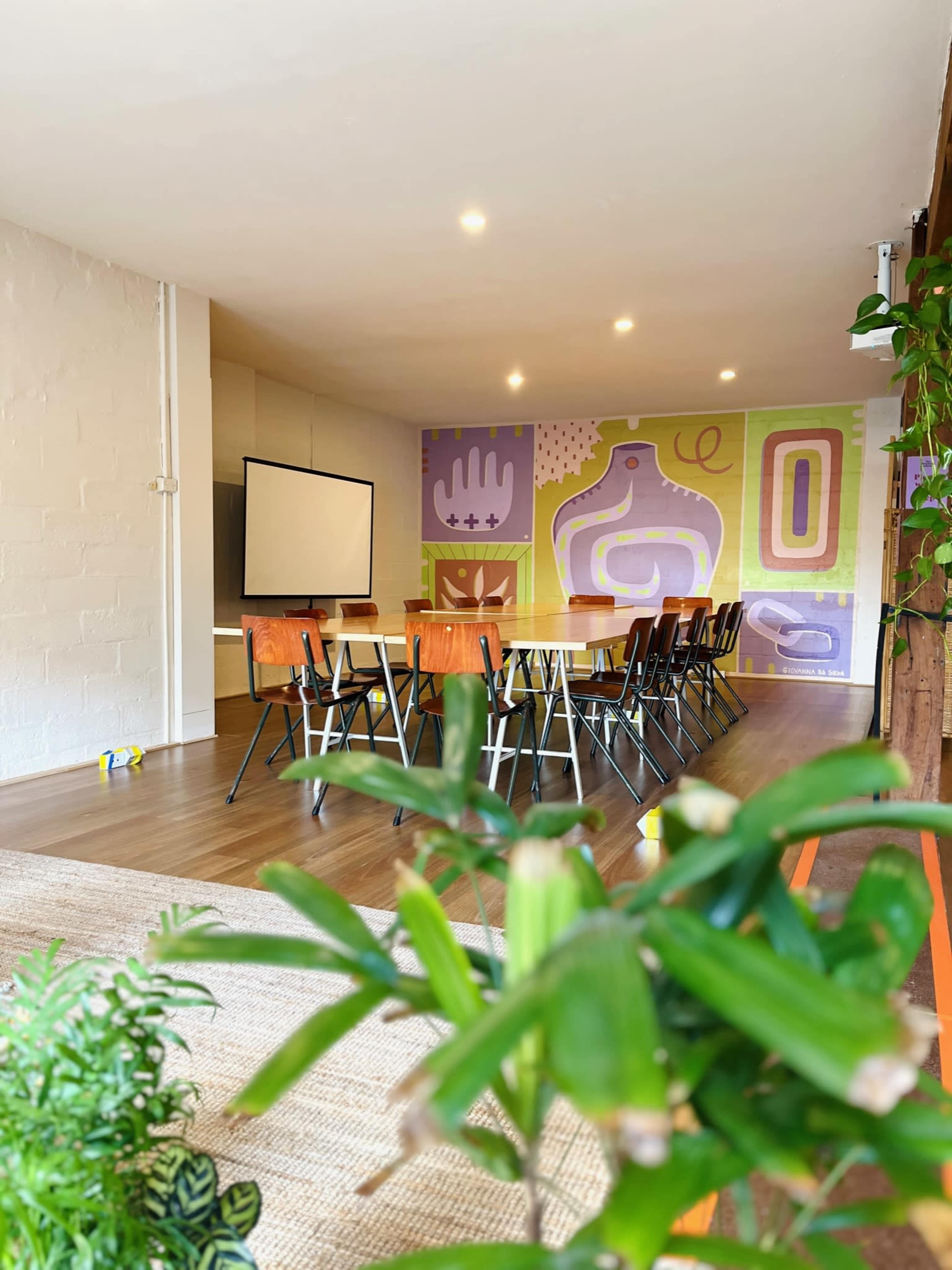 A spacious meeting room features a long table surrounded by chairs, with a colorful mural on the wall and a projector screen at the front.