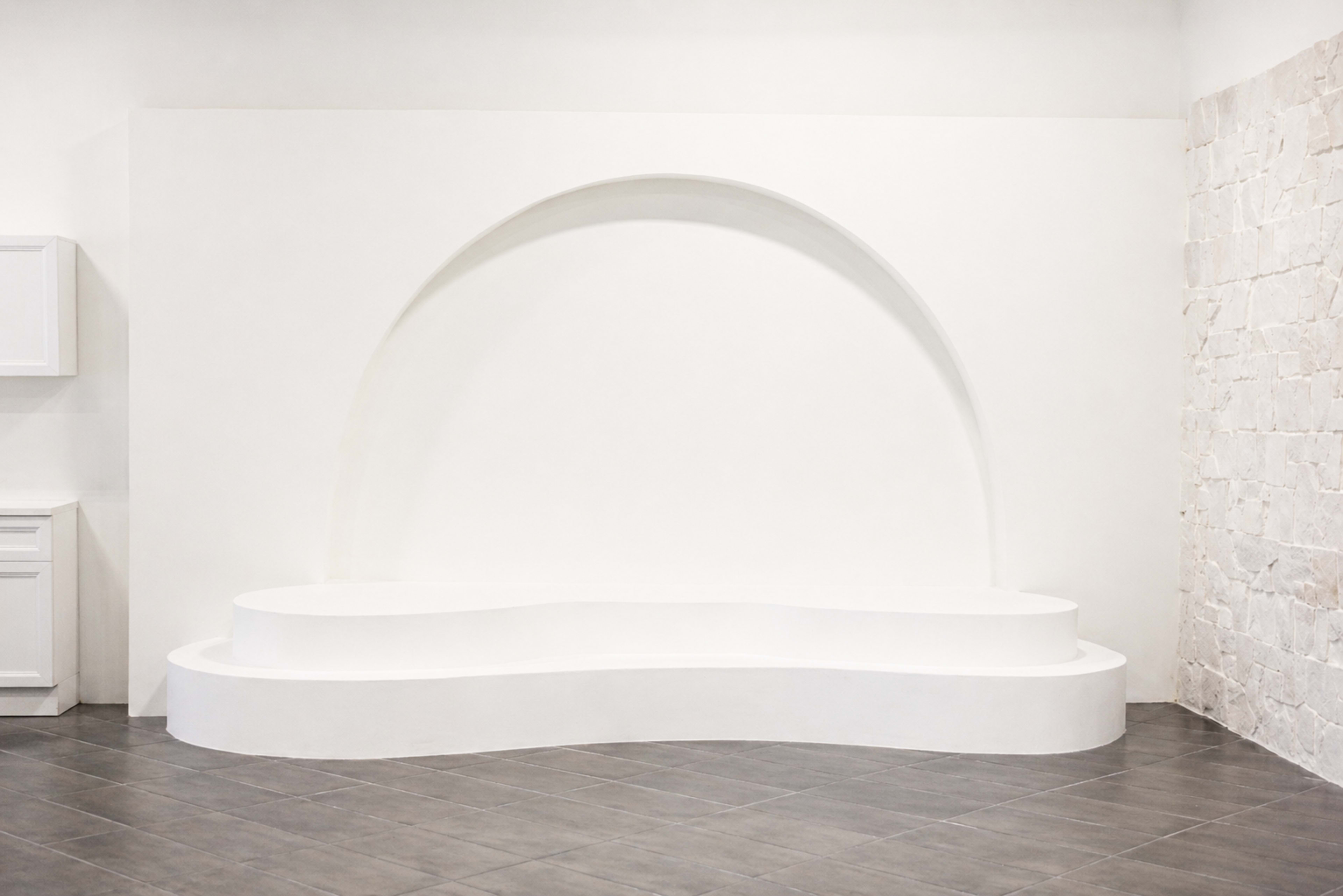 The image depicts a minimalist white wall with a large curved recess and a matching curved platform in a room with tiled flooring.