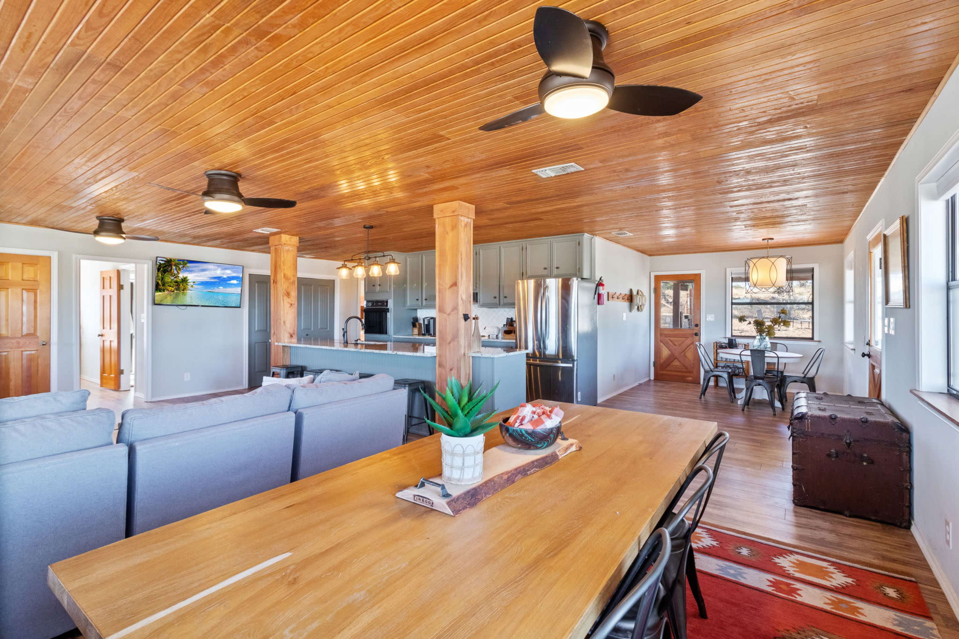 A modern open-concept living and dining area with wooden flooring, a large wooden dining table, a sectional sofa, and a kitchen visible in the background.