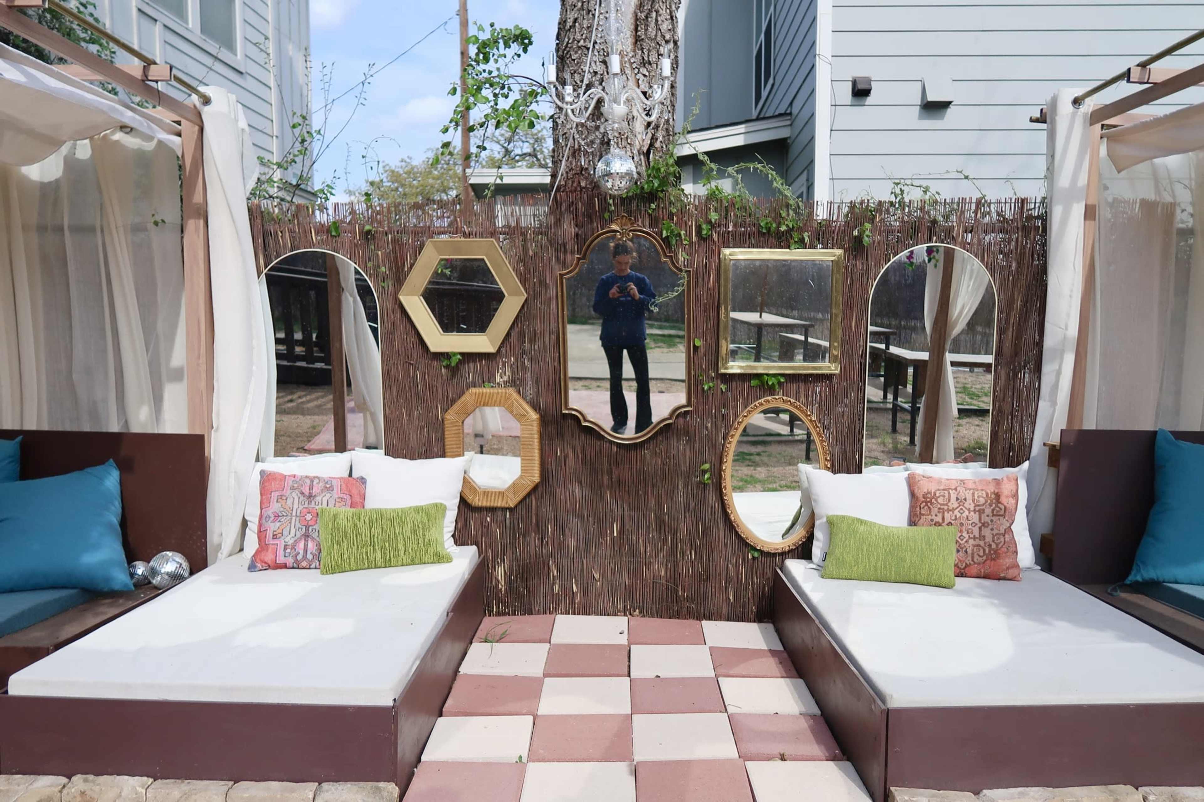 The image shows a cozy outdoor lounge area with cushioned seating, decorative mirrors on a bamboo backdrop, and a checkerboard flooring pattern.