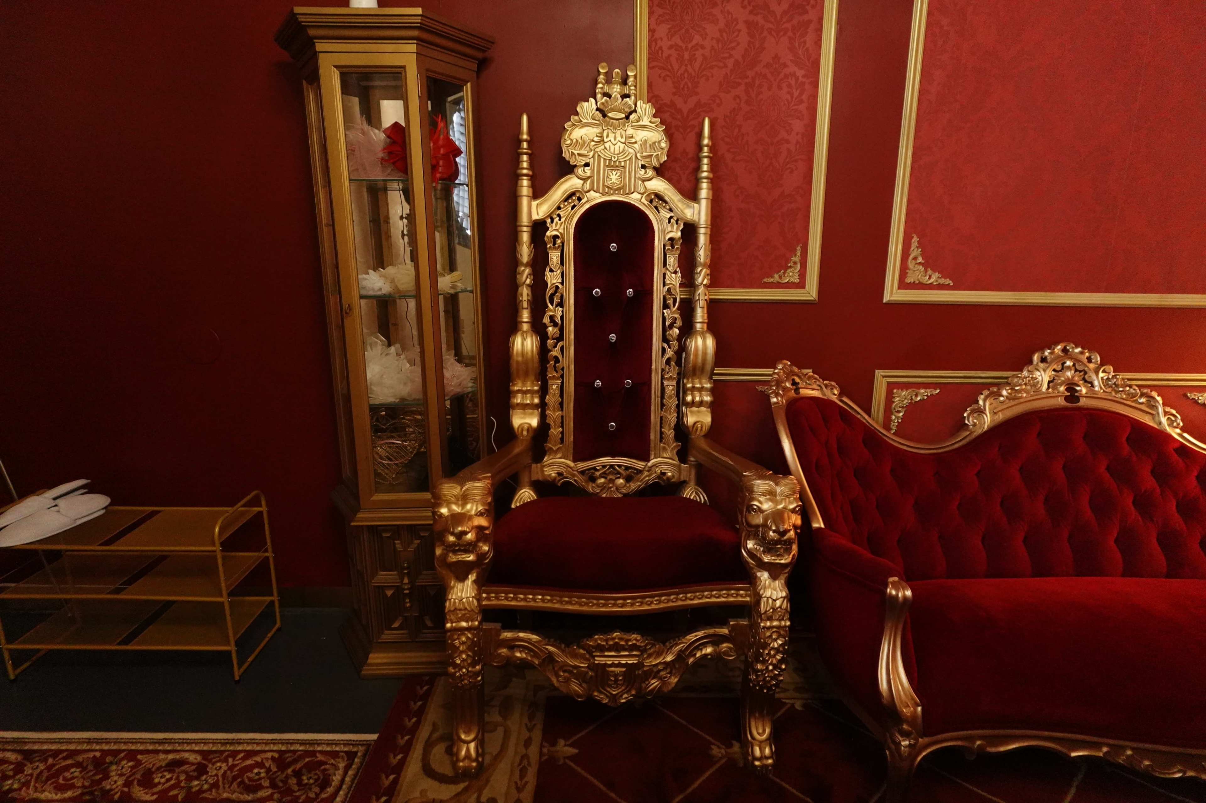 The image depicts an ornate golden throne with a red velvet seat, positioned against a deep red wallpapered wall, accompanied by a plush red sofa and a glass display cabinet.