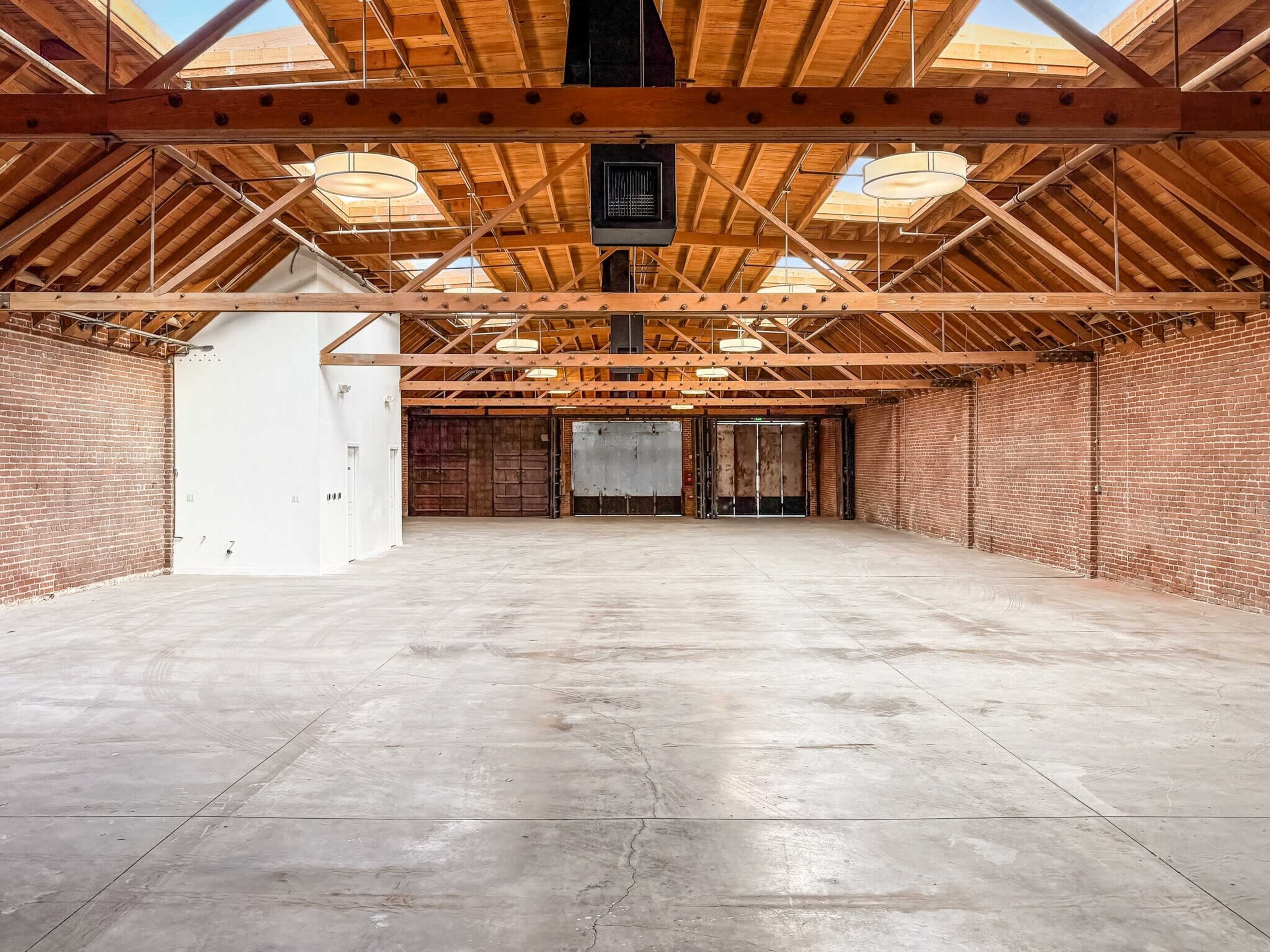The image shows a spacious, minimalistic interior of a room with exposed brick walls and a high wooden ceiling.