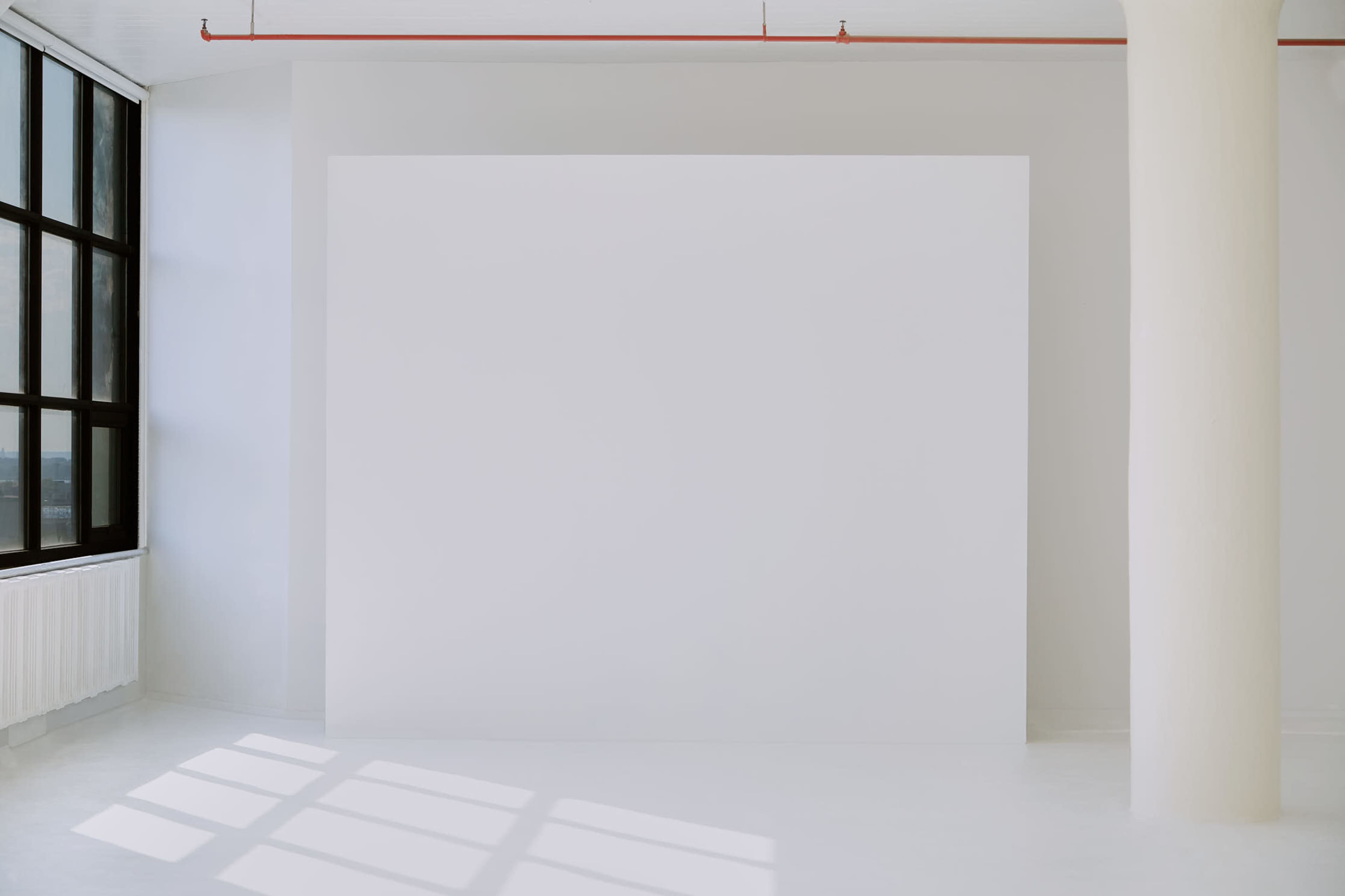 A bright, minimalistic interior space with a large blank white wall and a tall window.