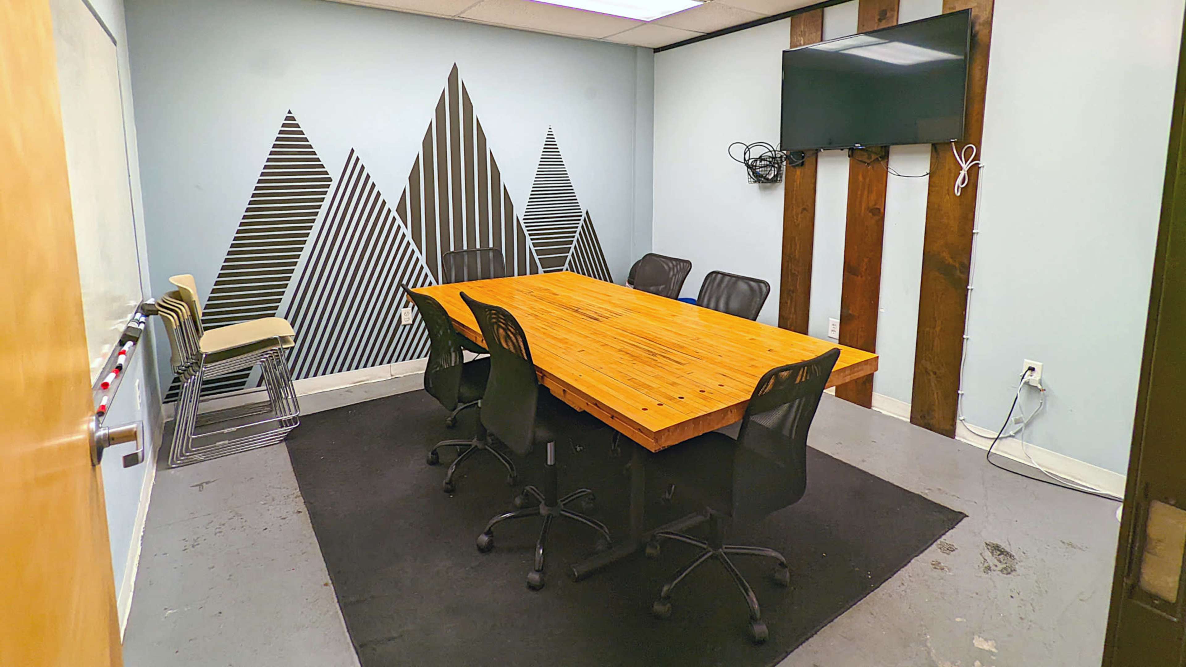 A conference room features a wooden table surrounded by six black chairs, with a wall displaying geometric patterned art and a television mounted above.