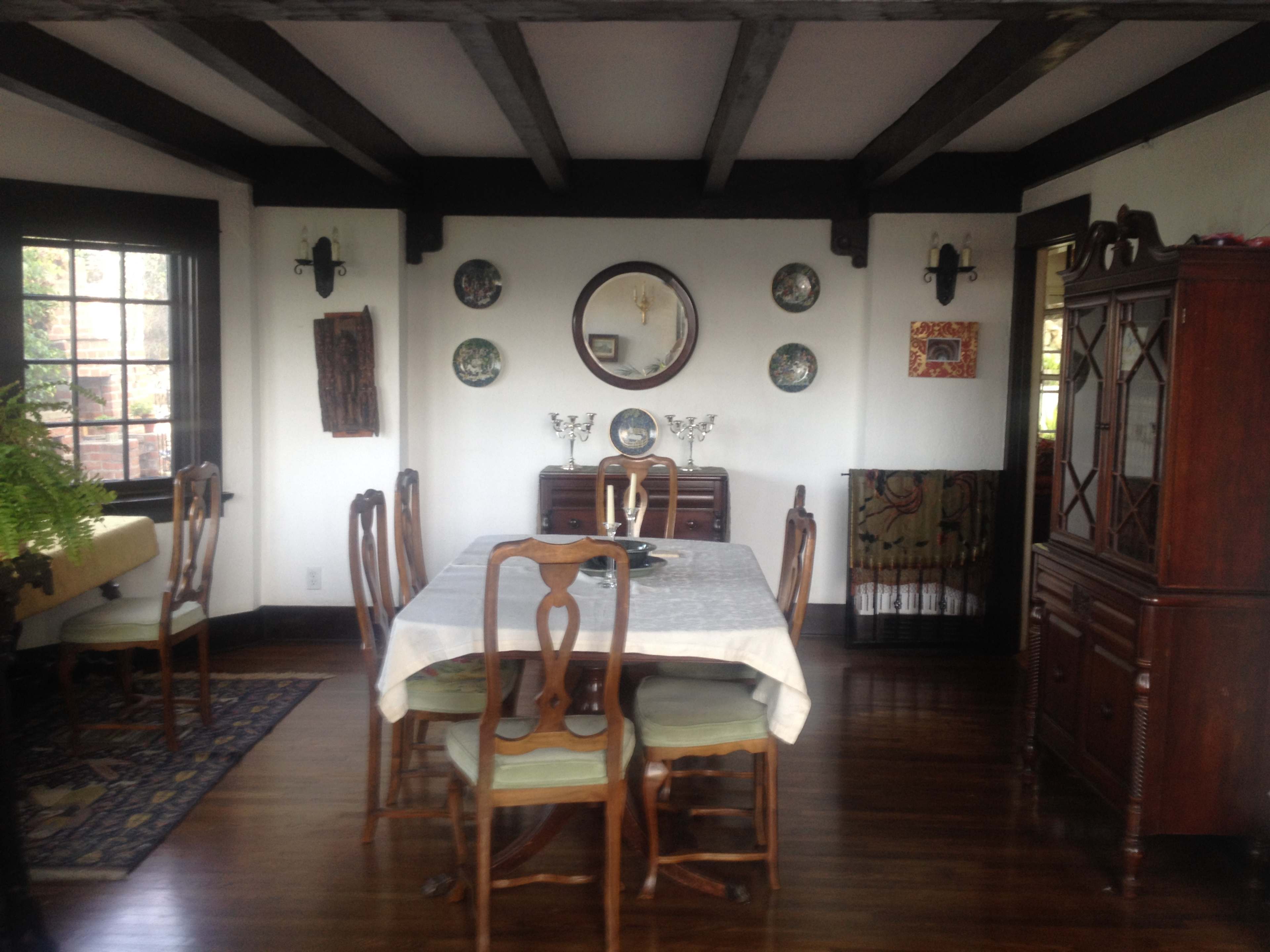 The image shows a dining room with a wooden table set for six, surrounded by antique chairs, and featuring a cabinet against the wall with decorative items.