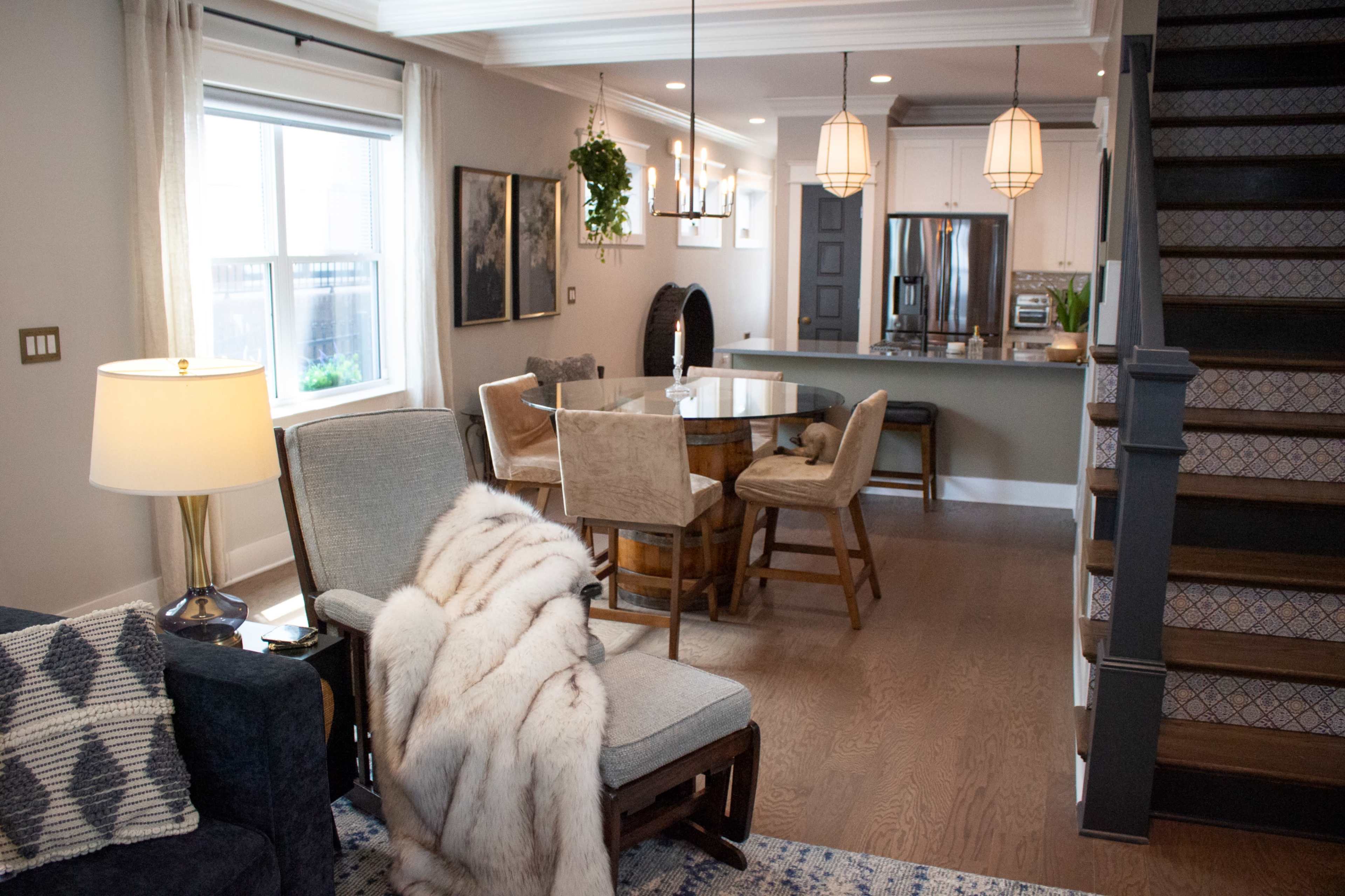 The image shows a stylish, modern living space featuring a dining area with a wooden table and chairs, a cozy seating area with a gray armchair draped with a blanket, and a staircase leading to the upper level.