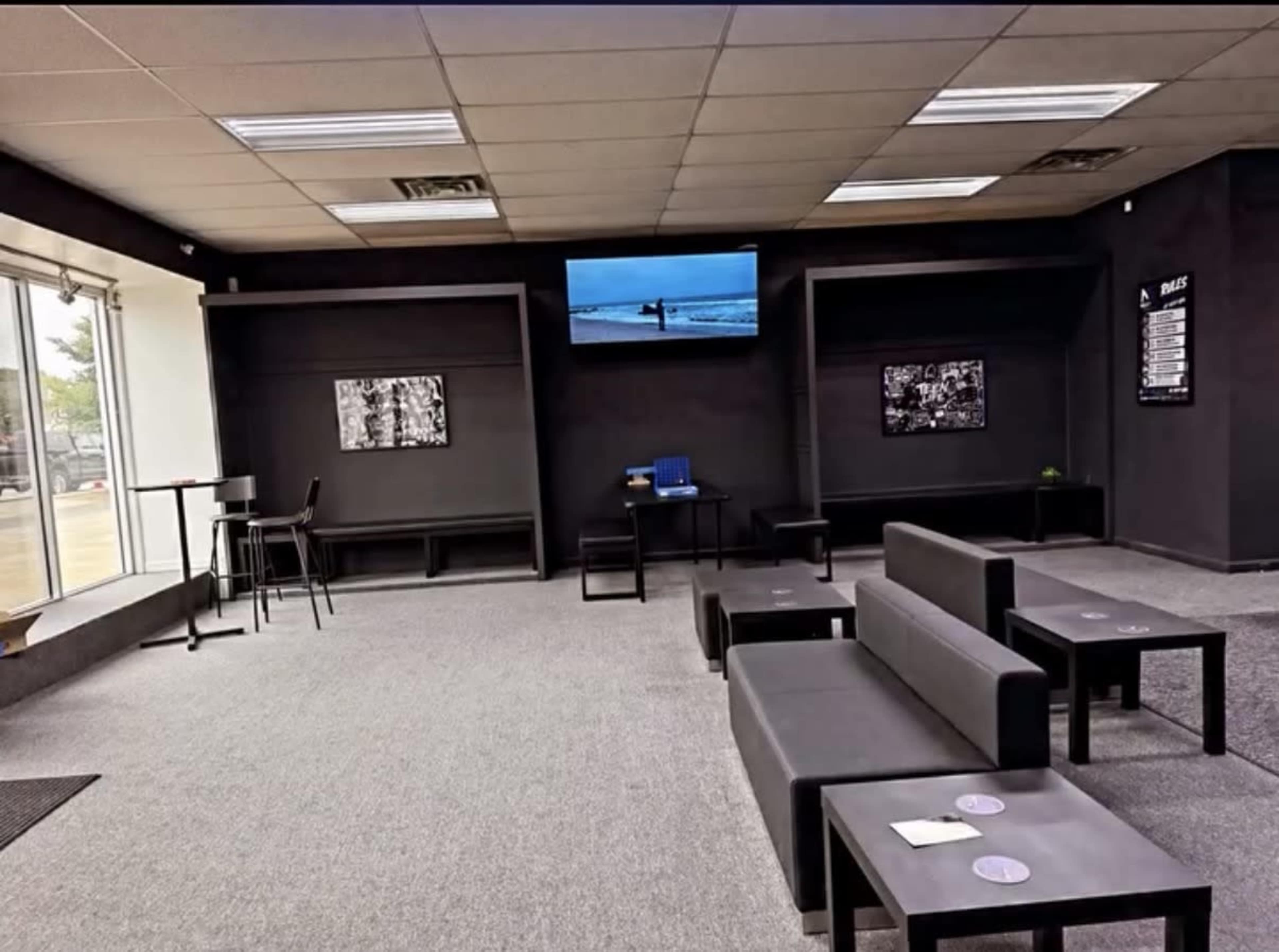 The image shows a modern interior of a lounge area with dark walls, several black couches, a small table, and a television mounted on the wall.