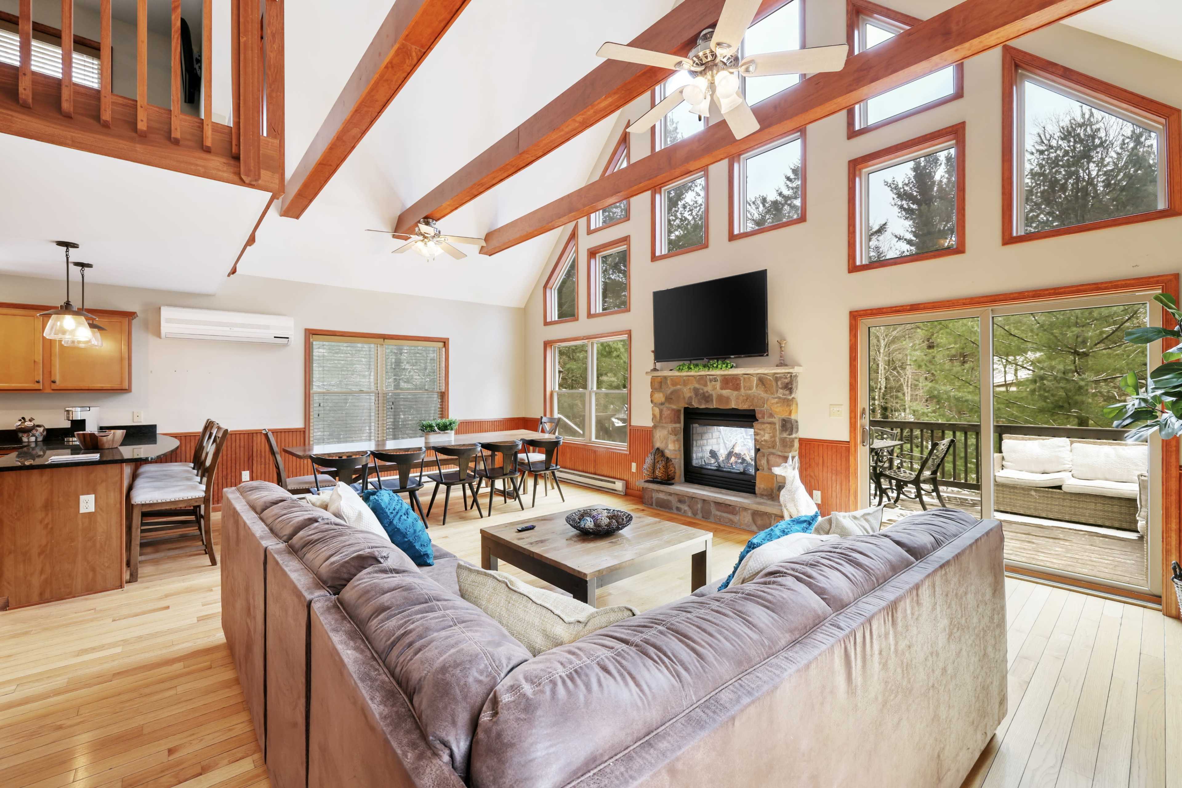 A spacious living room with large windows, a stone fireplace, and a comfortable sectional sofa, leading to a deck area with outdoor seating.