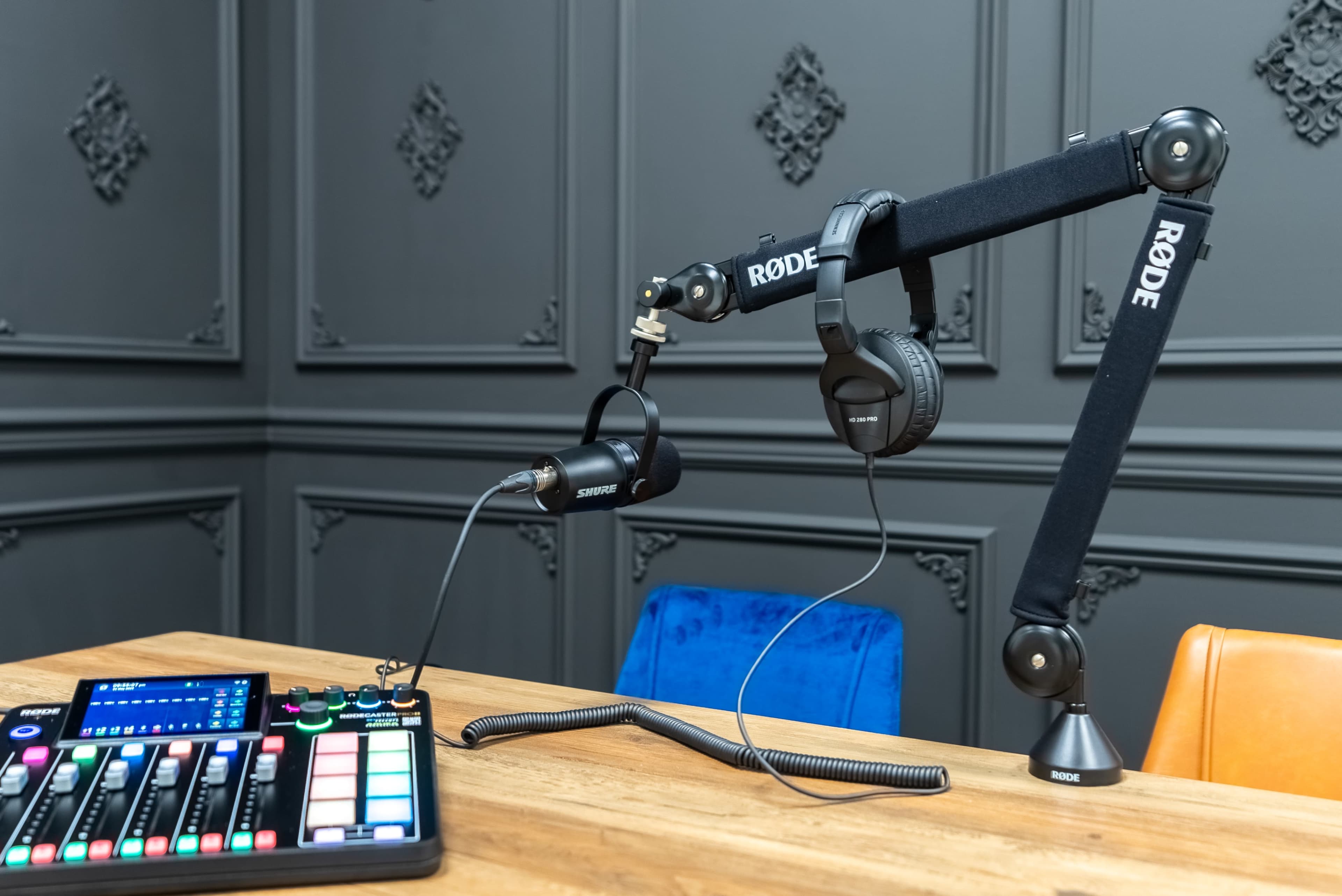 A podcasting setup features a microphone on an adjustable arm, headphones, and a control panel on a wooden table against a decorative gray wall.