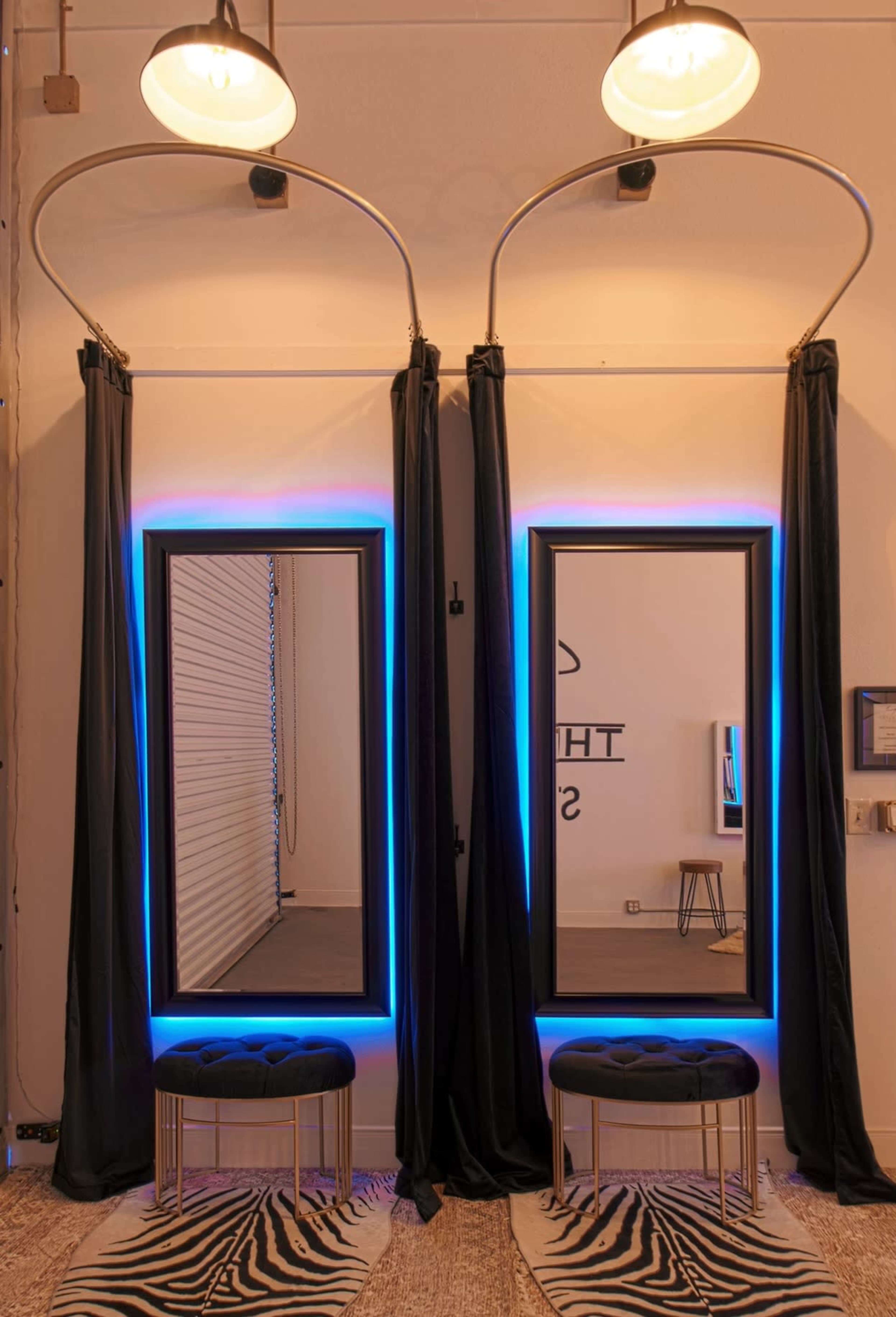 Two illuminated mirrors framed in black, flanked by dark curtains, with two upholstered benches in front on a patterned rug.