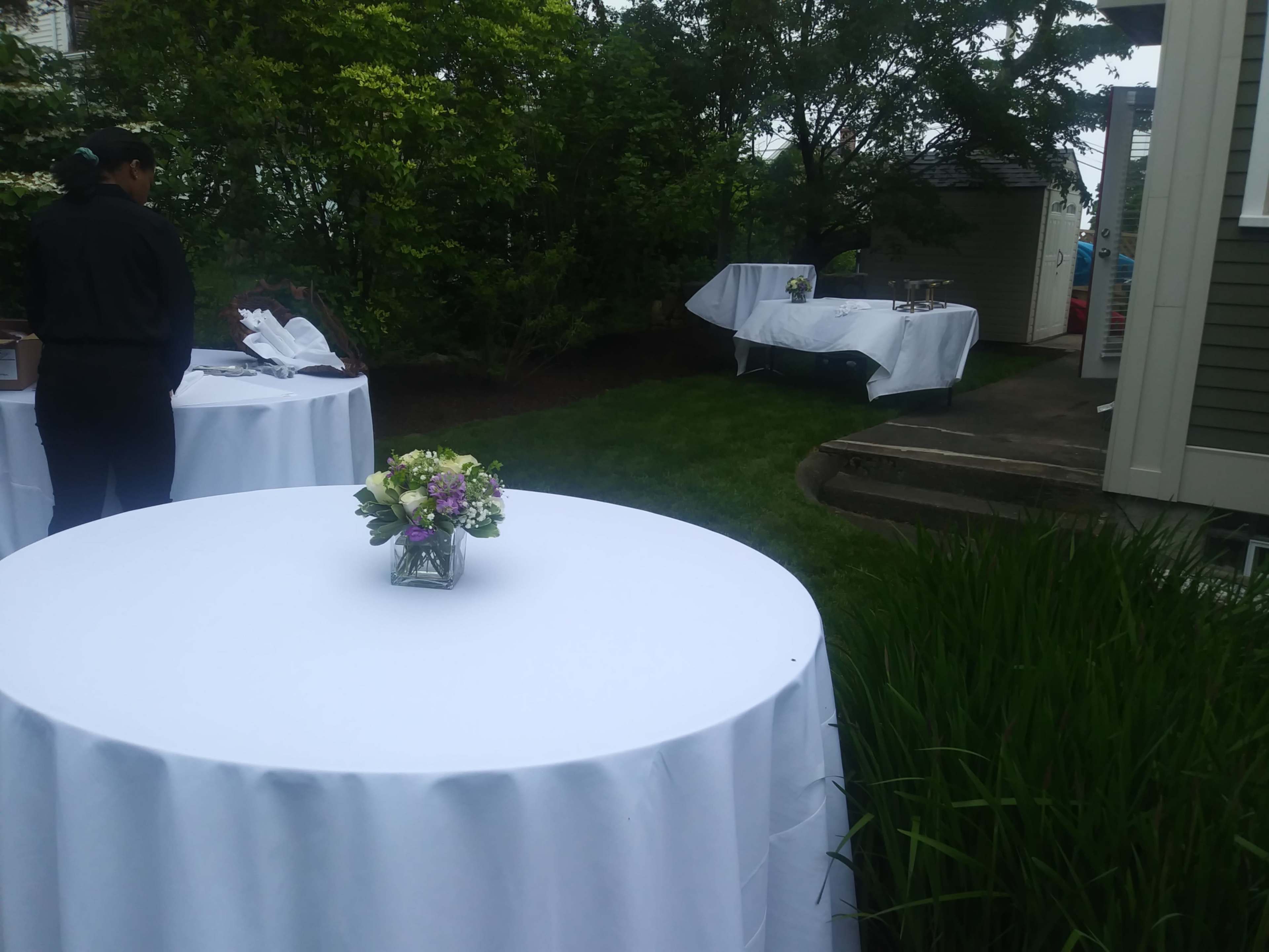 A garden area is set up for an event with white tables covered by tablecloths and a floral centerpiece on each table.