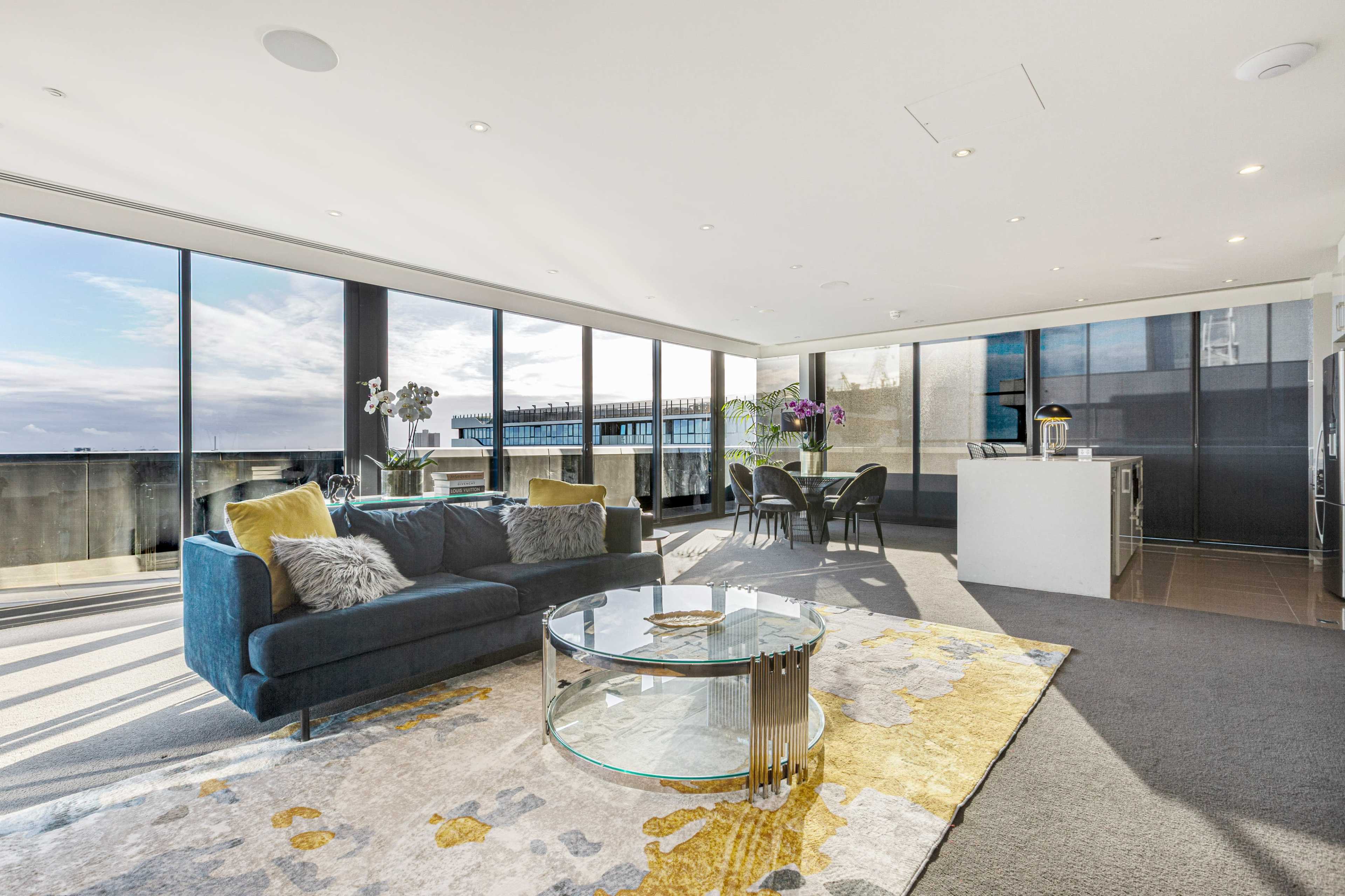 The image shows a modern living room with large floor-to-ceiling windows, a blue sofa, a circular glass coffee table, and a dining area with dark chairs and an open kitchen in the background.
