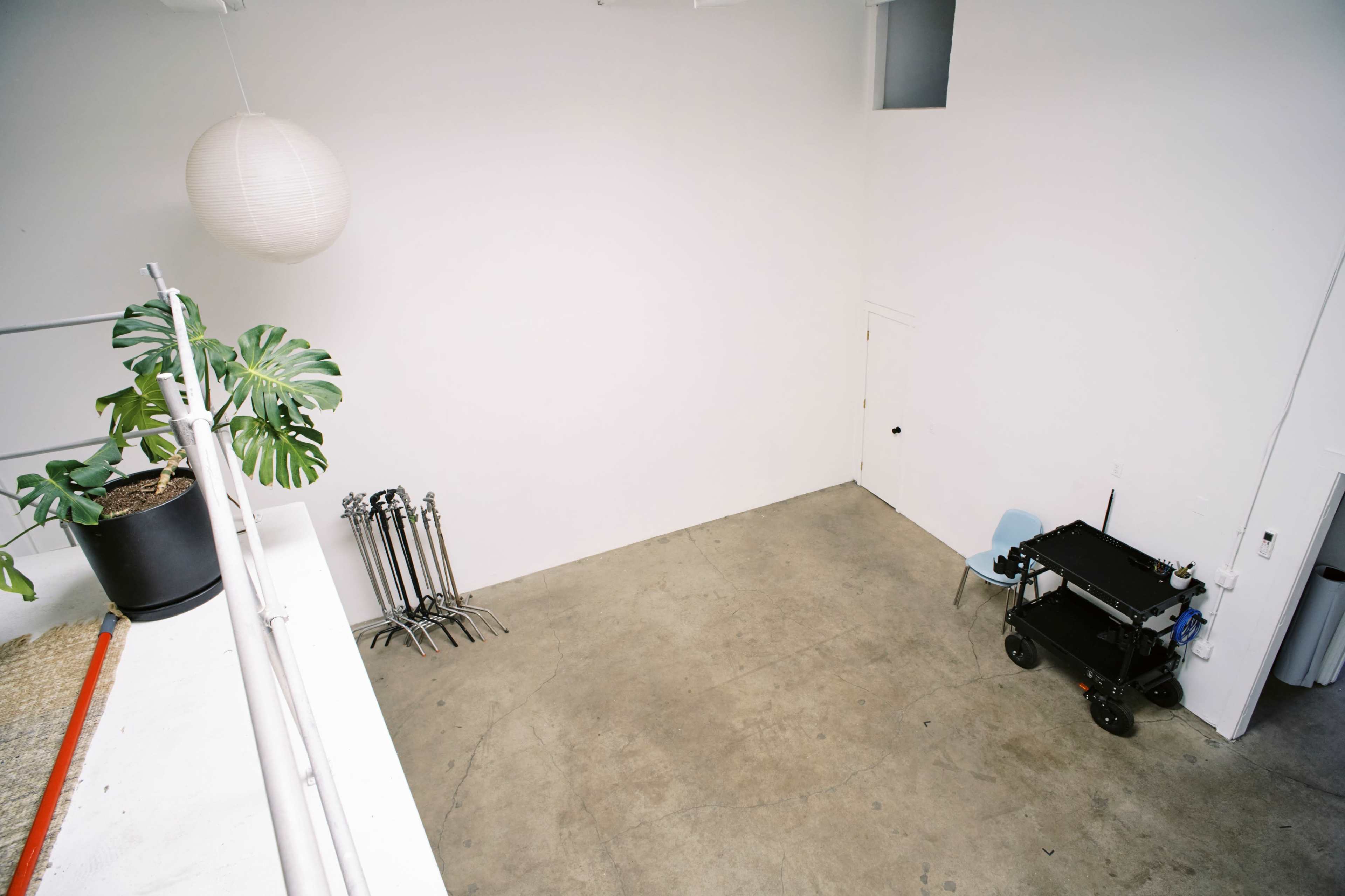 The image shows a spacious, empty room with concrete flooring, a small blue chair, a black cart, a plant in a pot, and several metal poles against the wall.