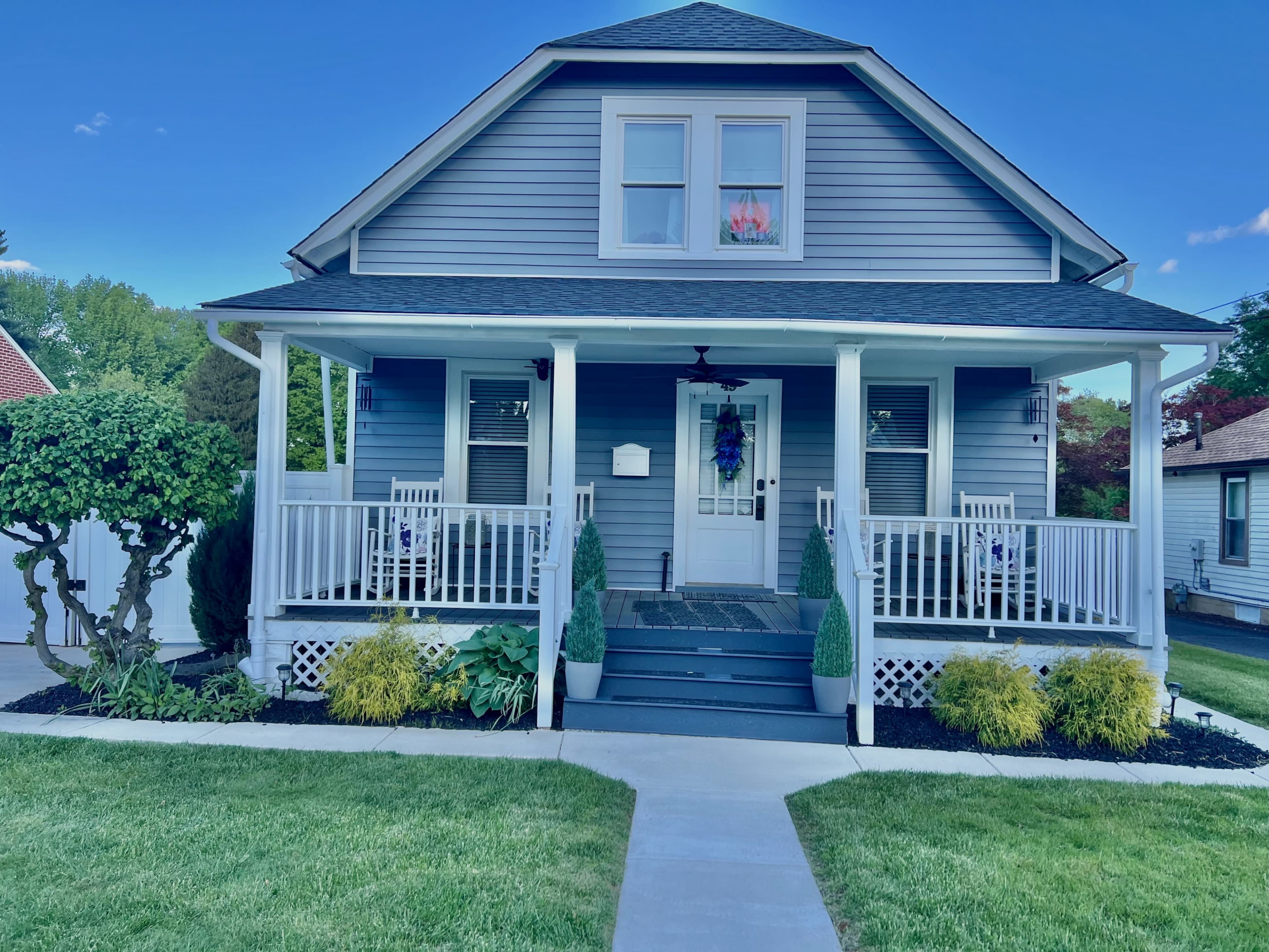 A two-story gray house with a front porch, white railings, and a well-maintained lawn features decorative plants and seating.