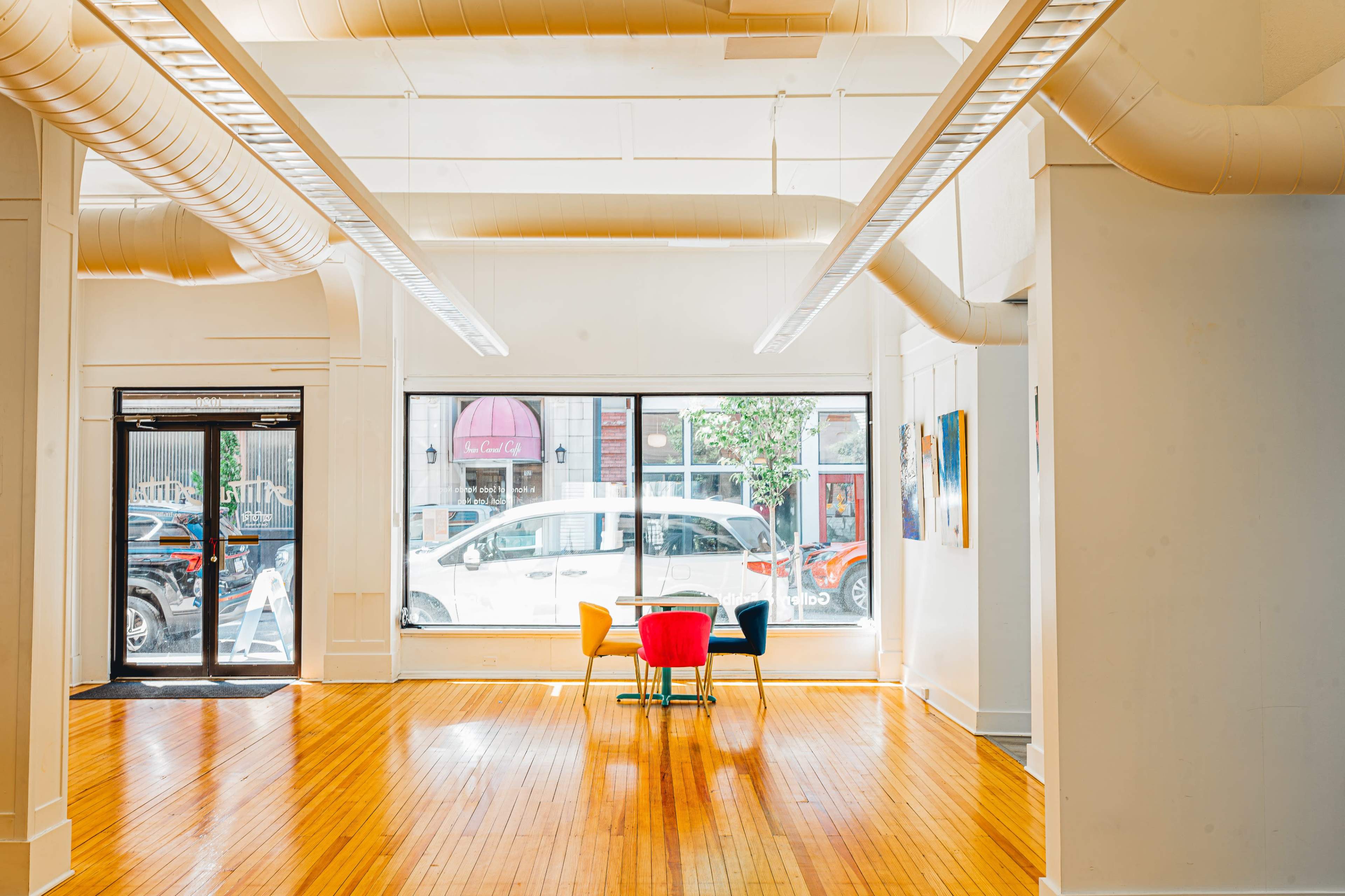 The image shows a spacious, brightly lit room with wooden flooring, featuring a small round table surrounded by colorful chairs near a large window.