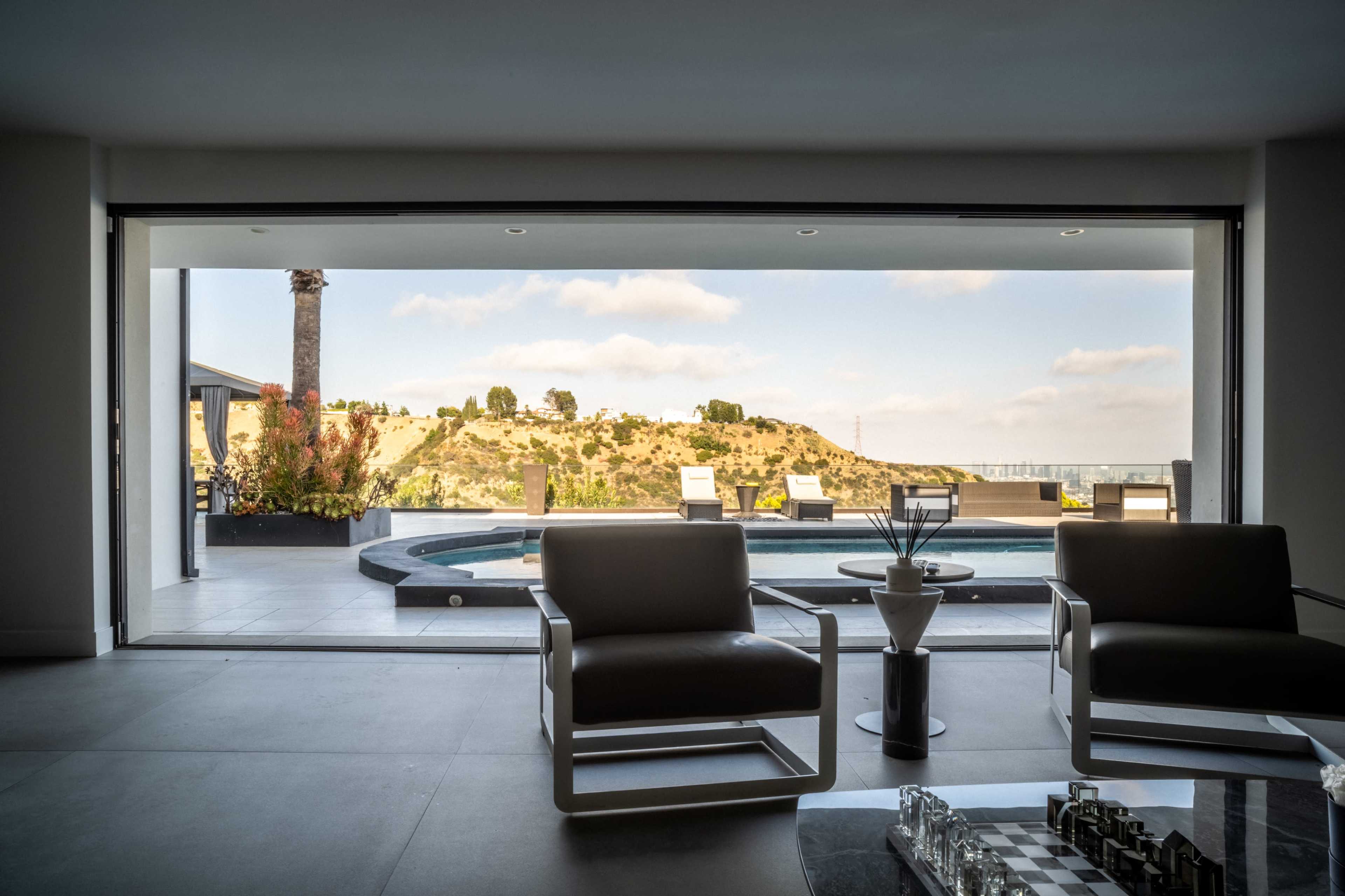 The image showcases a modern living room with large sliding glass doors that open to a scenic outdoor area featuring a pool, surrounded by hills and a clear blue sky.