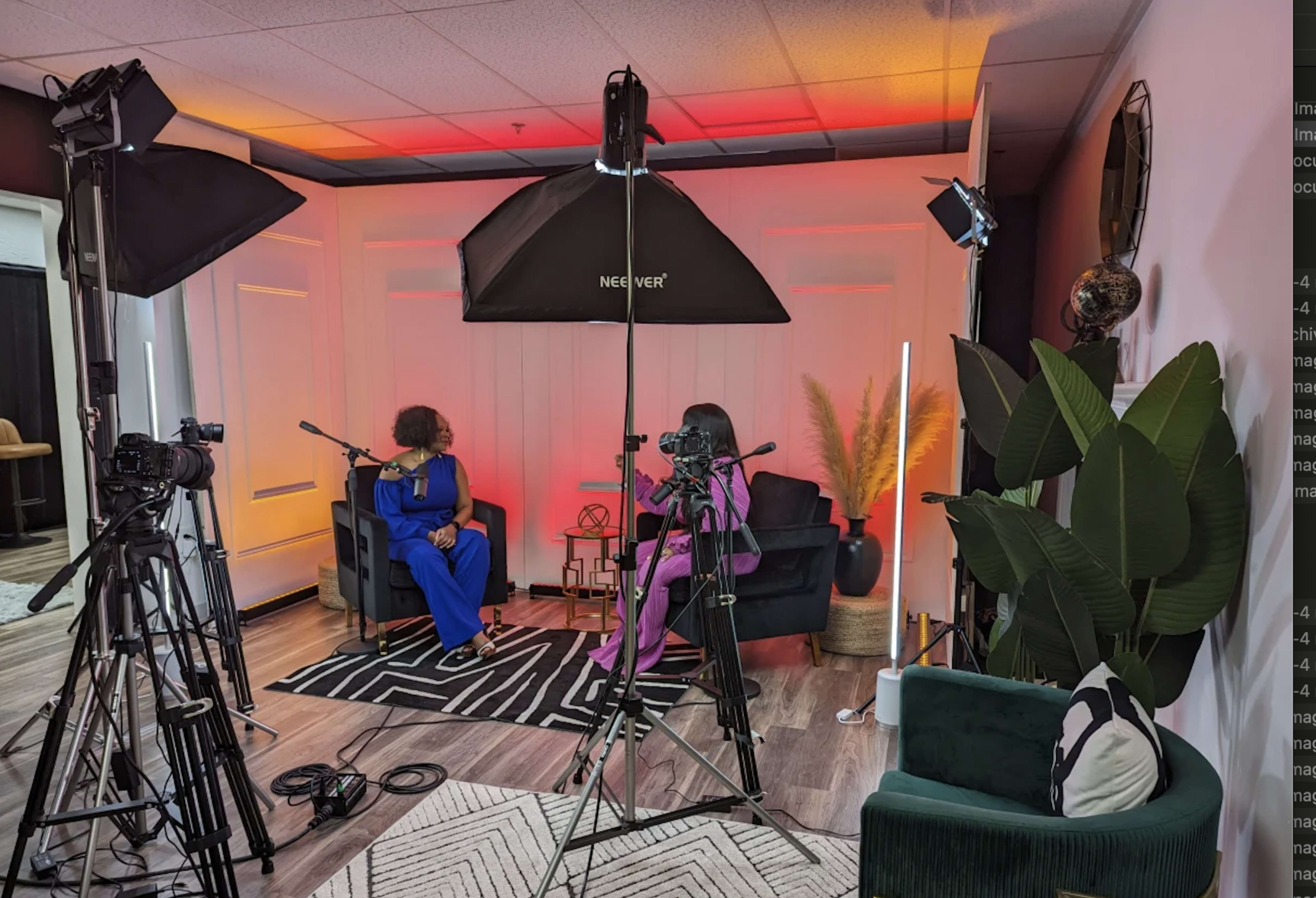A filming setup with two people seated in chairs, surrounded by video cameras and lighting equipment in a studio decorated with plants and lighting on the walls.