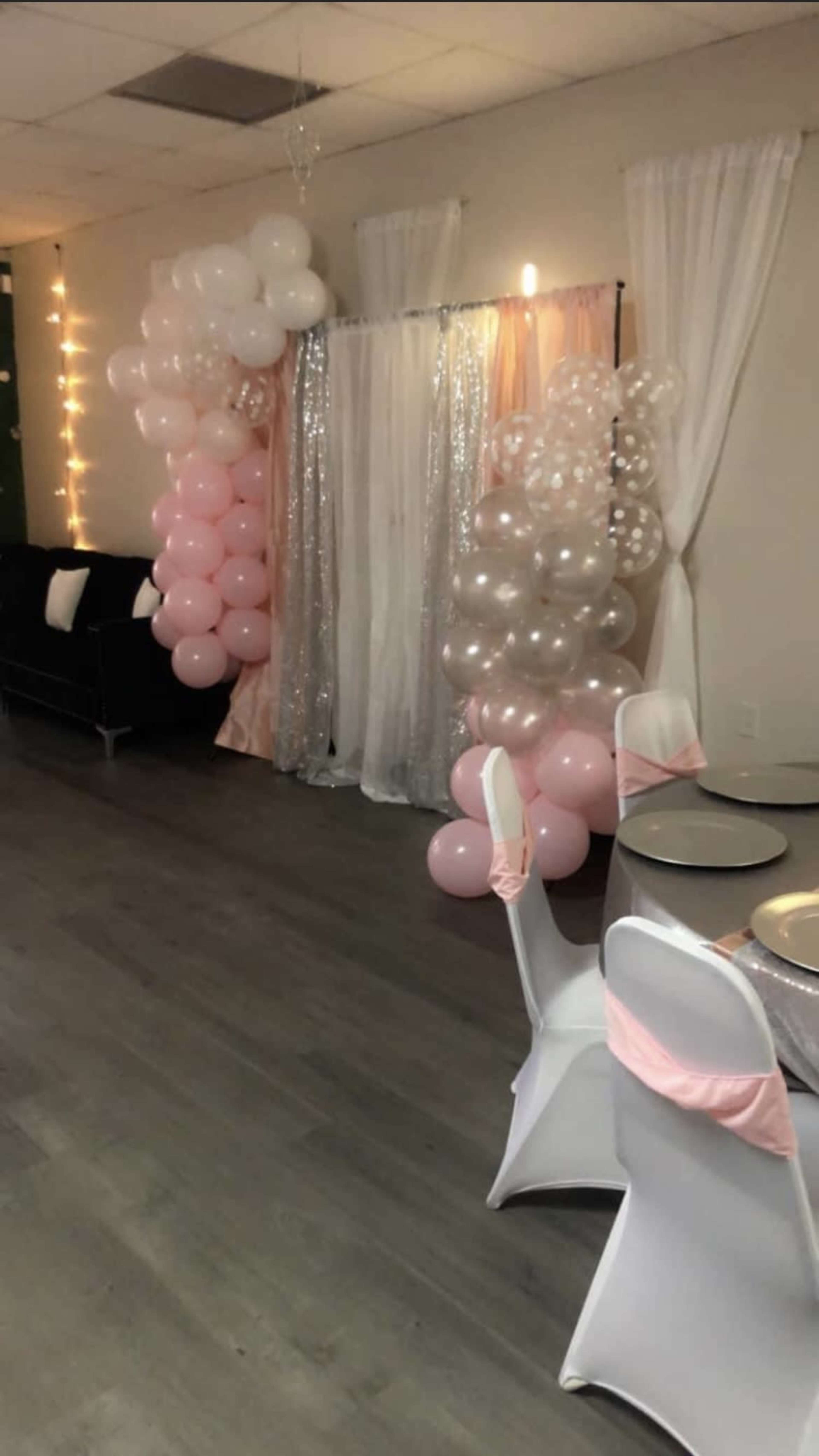 A decorated event space featuring balloon arrangements in shades of pink and silver, along with draped fabric backdrops and tables set with white chairs featuring pink sashes.