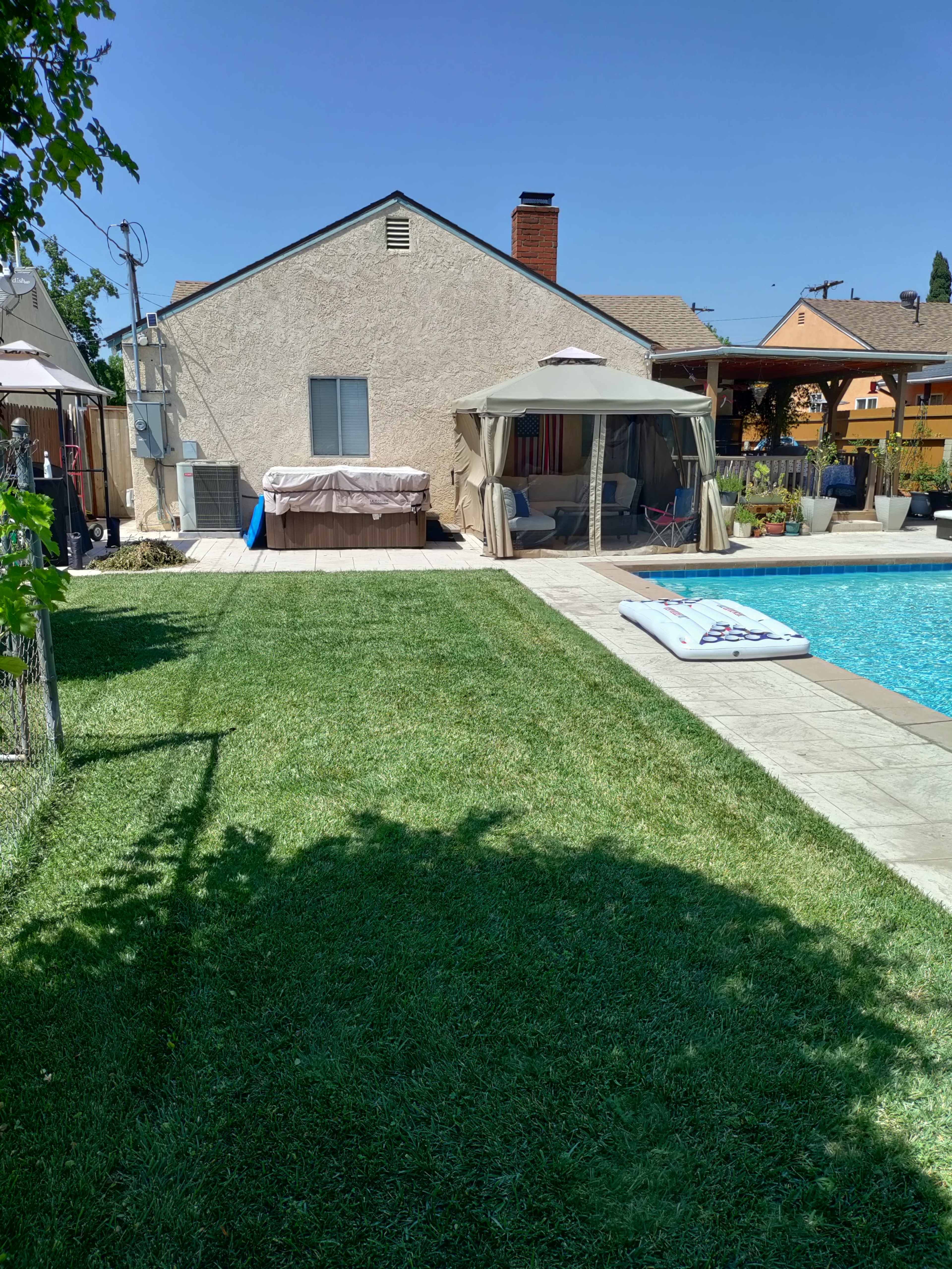 The image shows a backyard with a swimming pool, a gazebo, a hot tub, and a well-maintained lawn adjacent to a house.