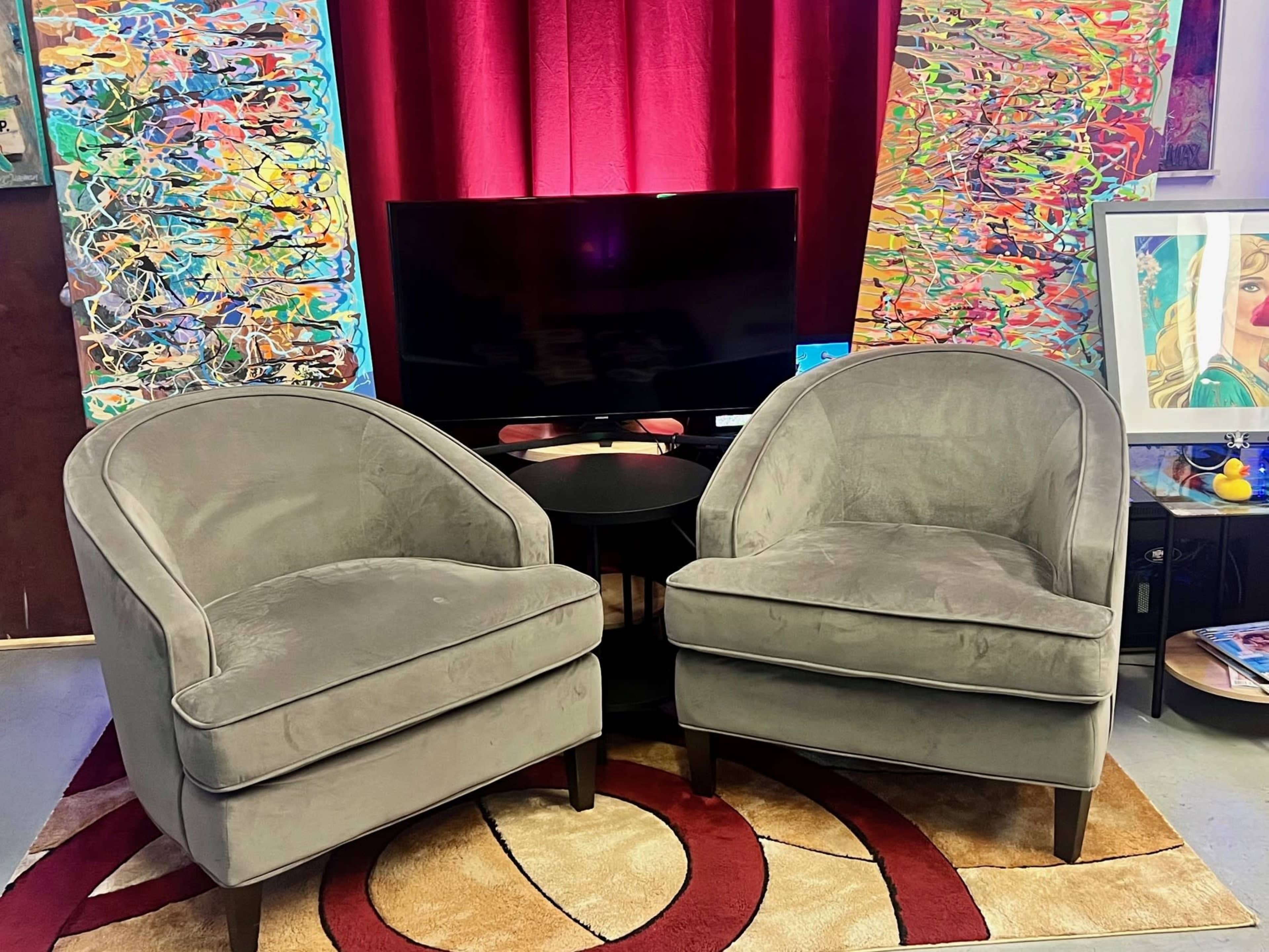 Two gray armchairs facing a black television, situated on a patterned rug, with colorful abstract artwork in the background.