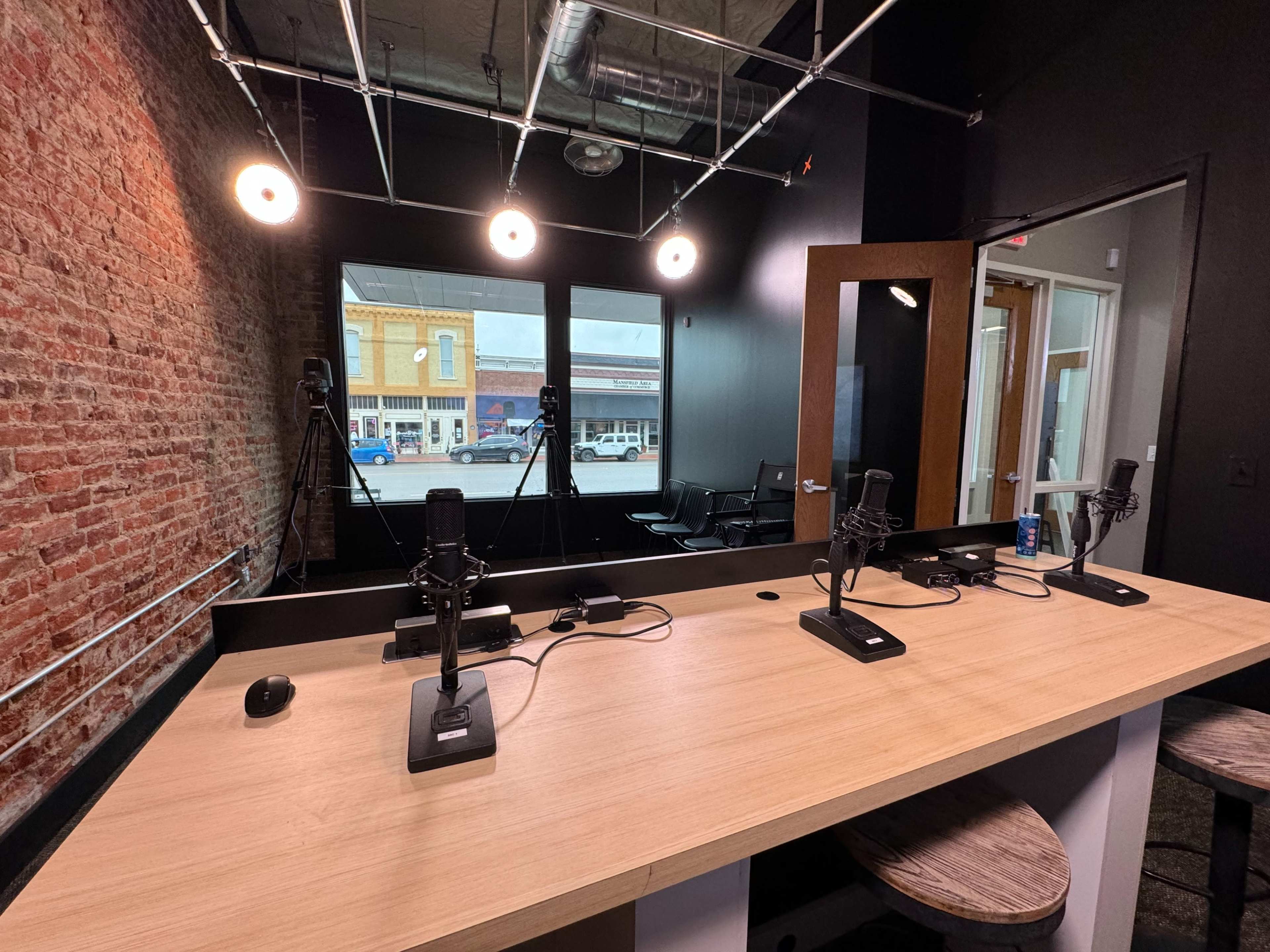 A podcast recording studio featuring a wooden table with microphones, chairs, and large windows overlooking a street.