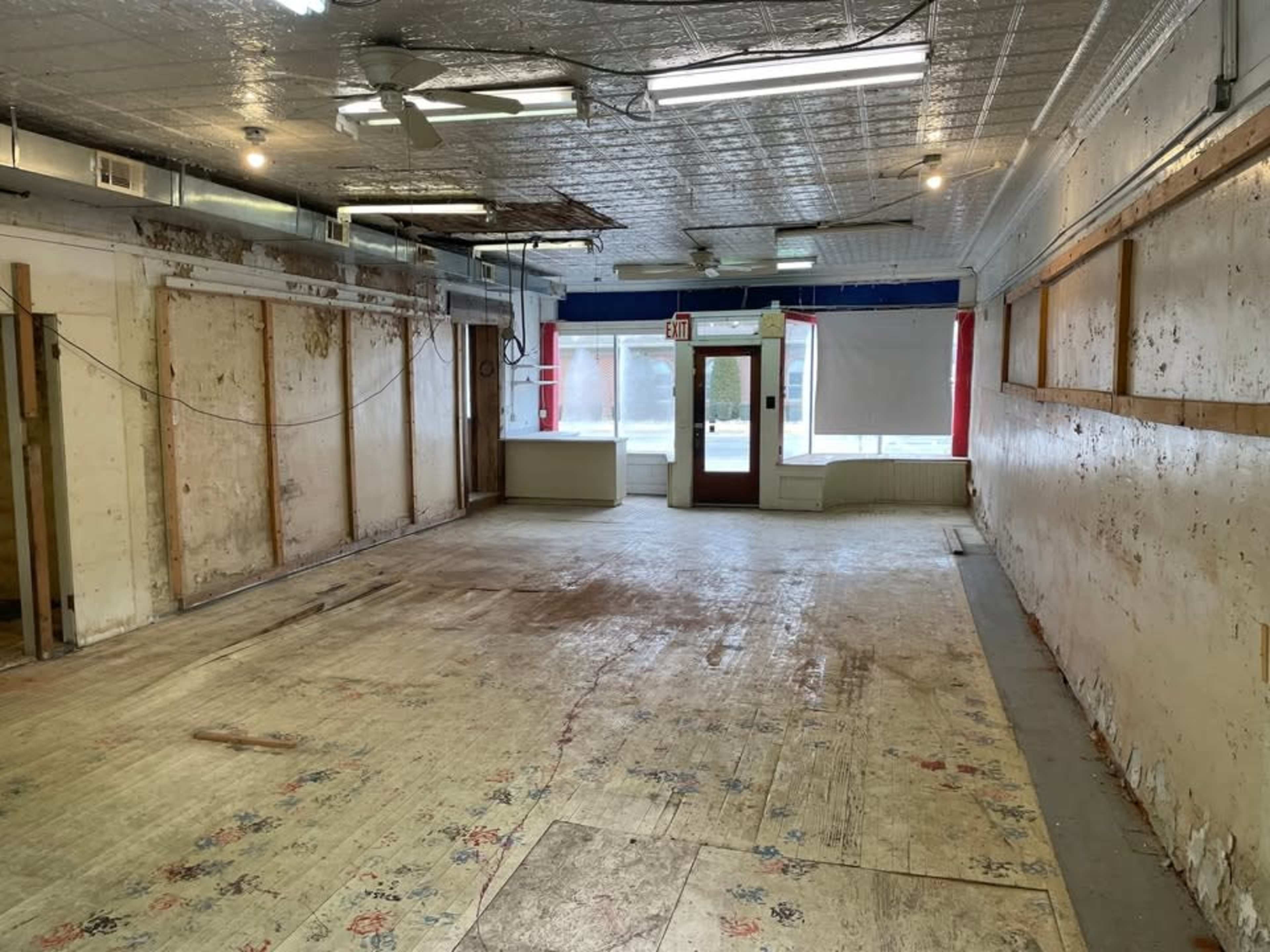 The image shows an empty, unfinished interior space with bare walls, a wooden floor, and large windows letting in natural light.