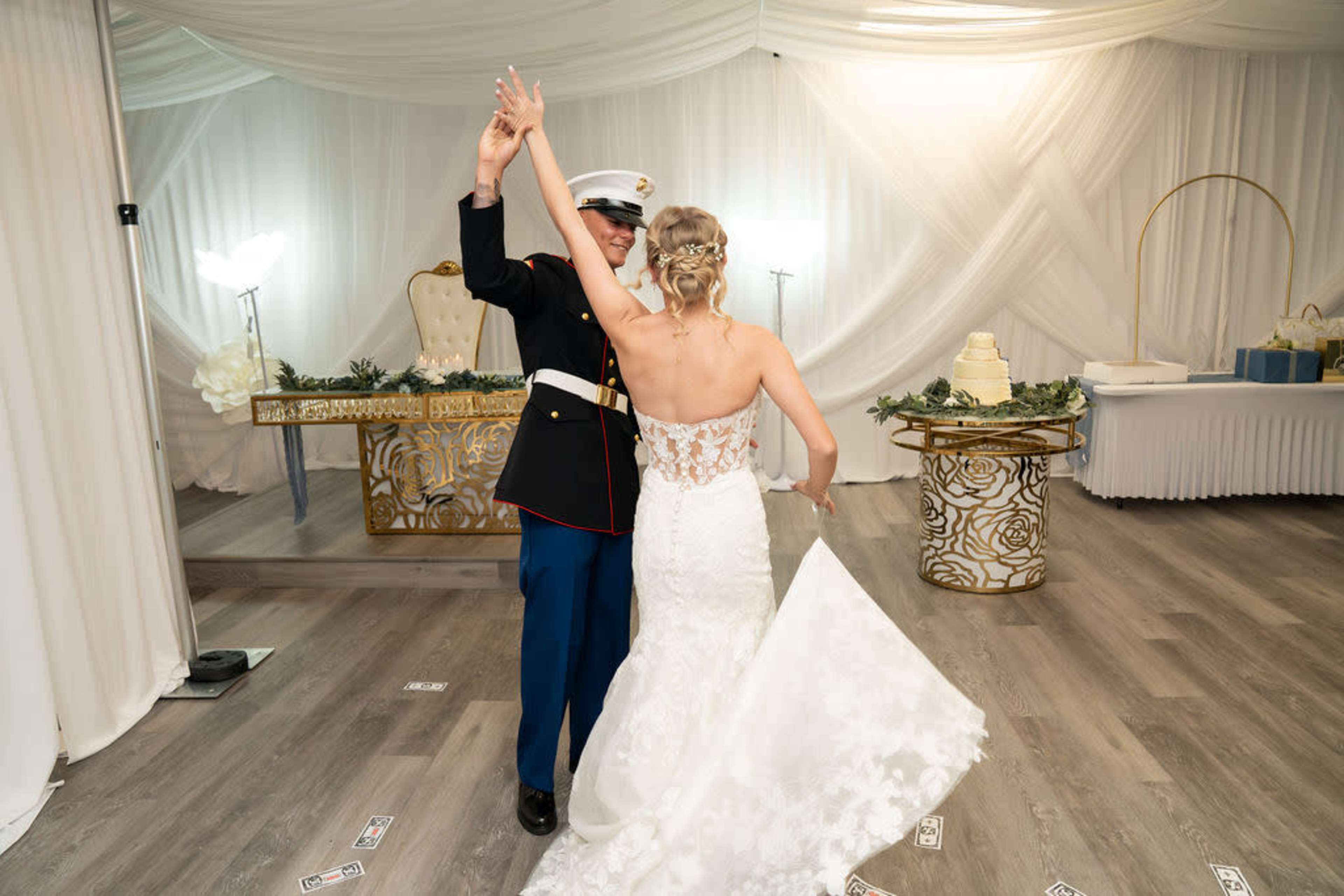 A bride in a lace wedding dress and a Marine in uniform dance together in a decorated reception hall.