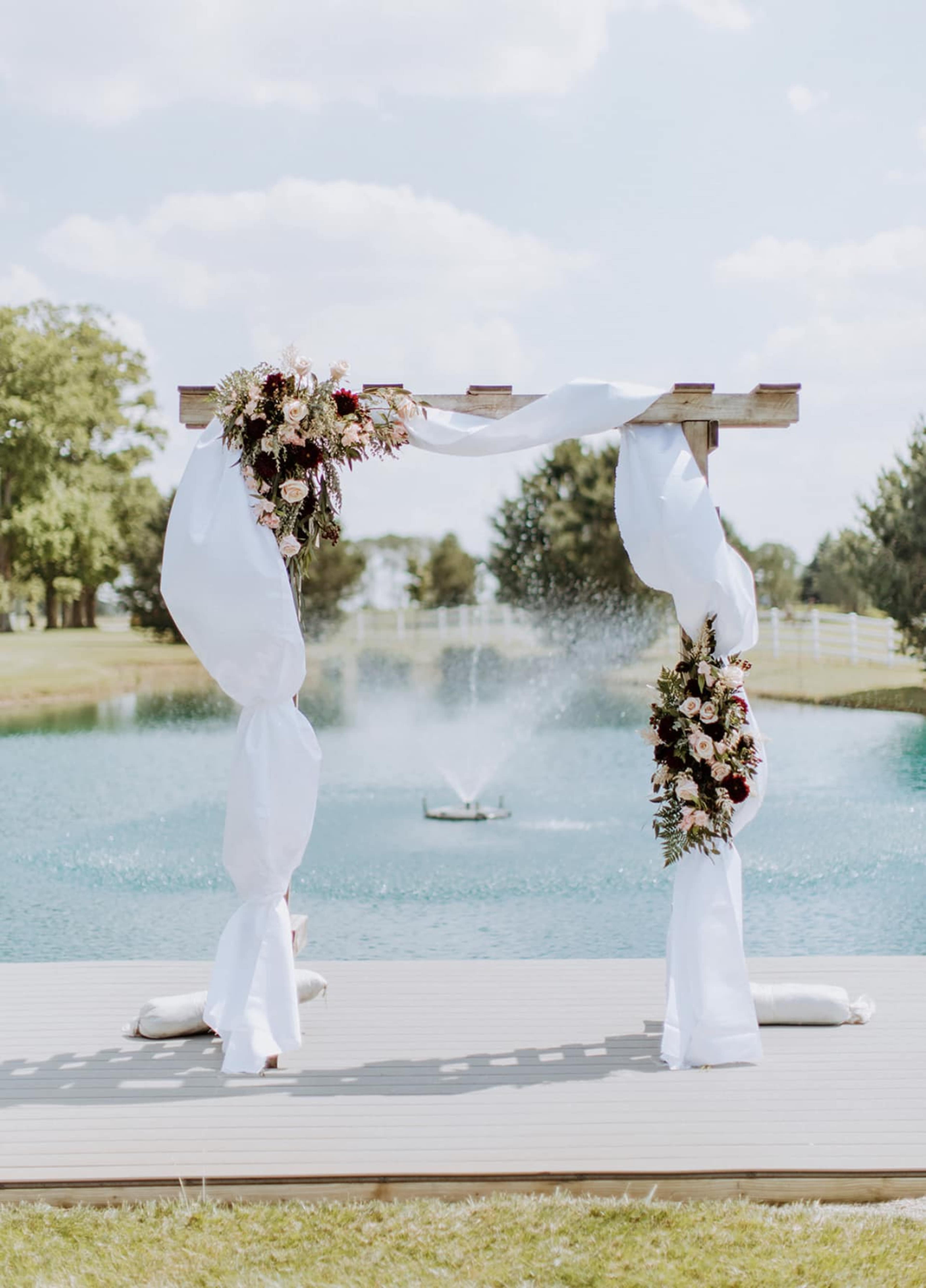 A wooden arch adorned with floral arrangements and draped fabric stands by a pond with a fountain in the background.