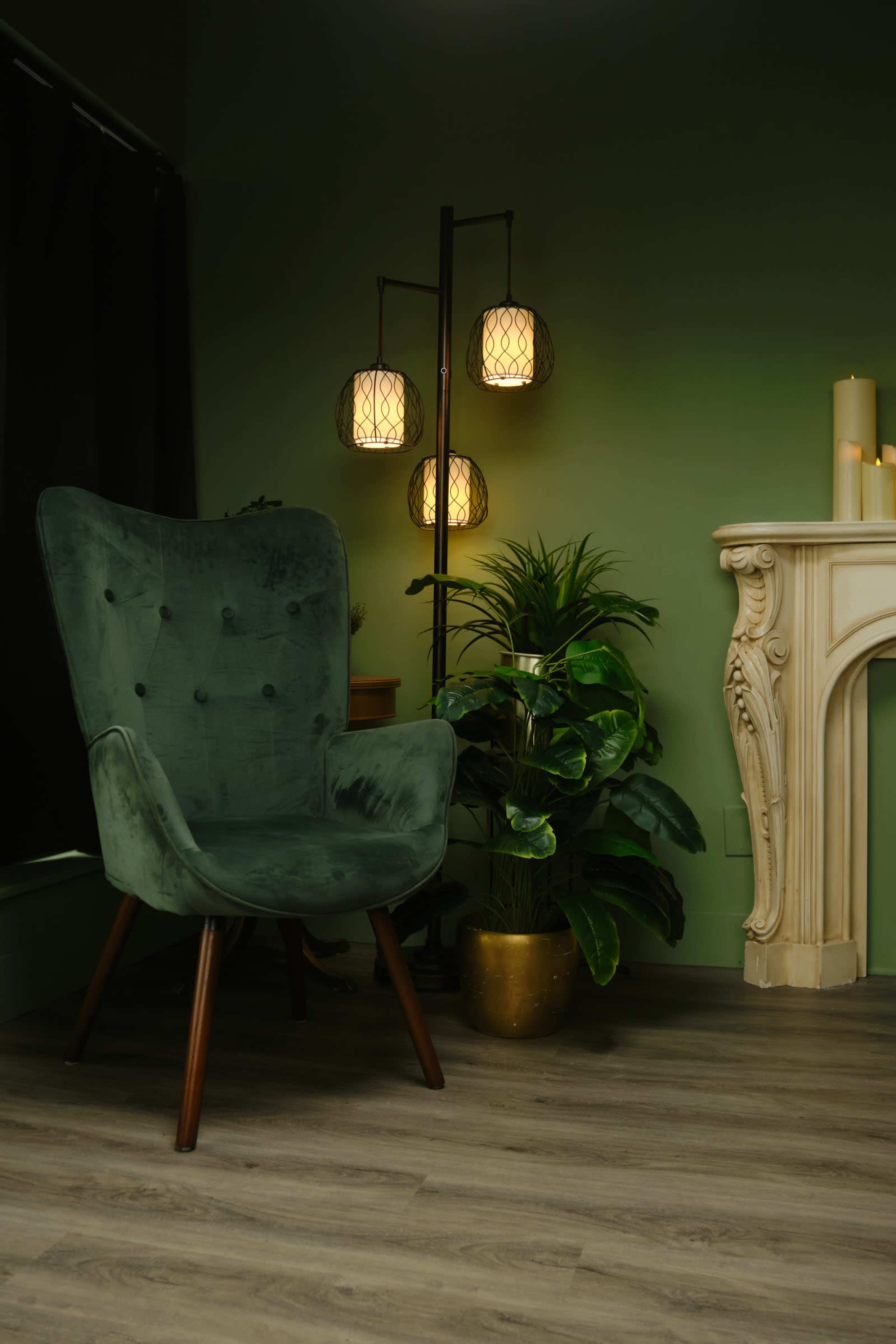 The image features a green velvet chair beside a decorative floor lamp and a potted plant, set against a muted green wall.