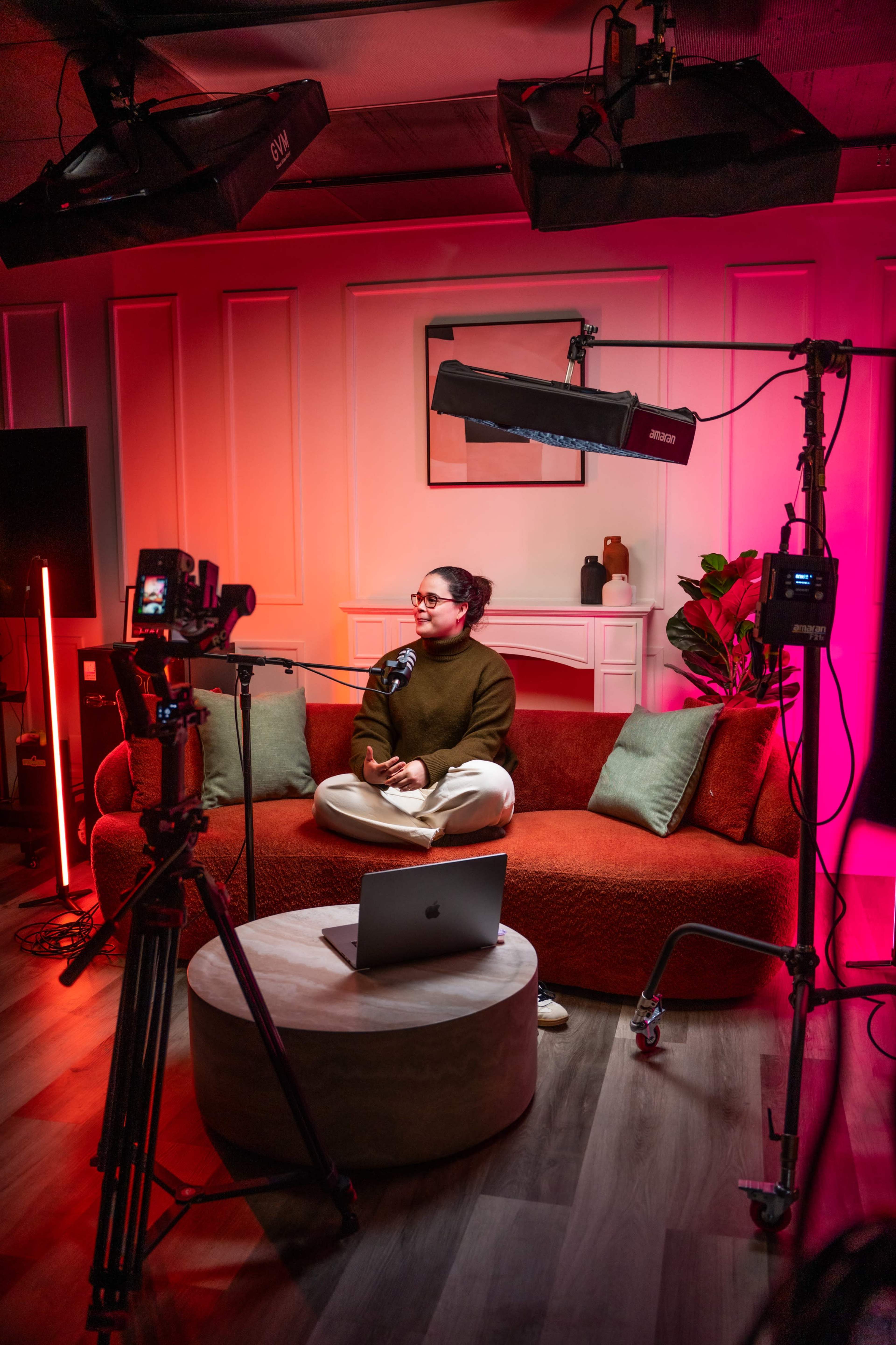 A person sits on a couch in a studio setup with cameras and lighting equipment, preparing to record or broadcast content.