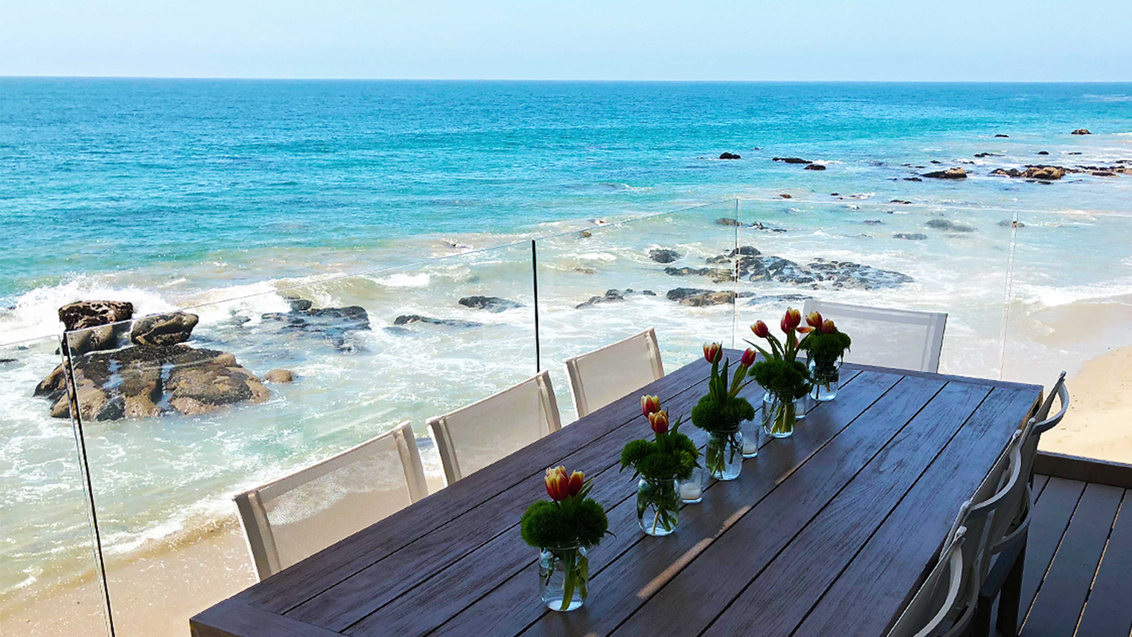 A seaside table is set with vases of flowers, overlooking the ocean and rocky shore.