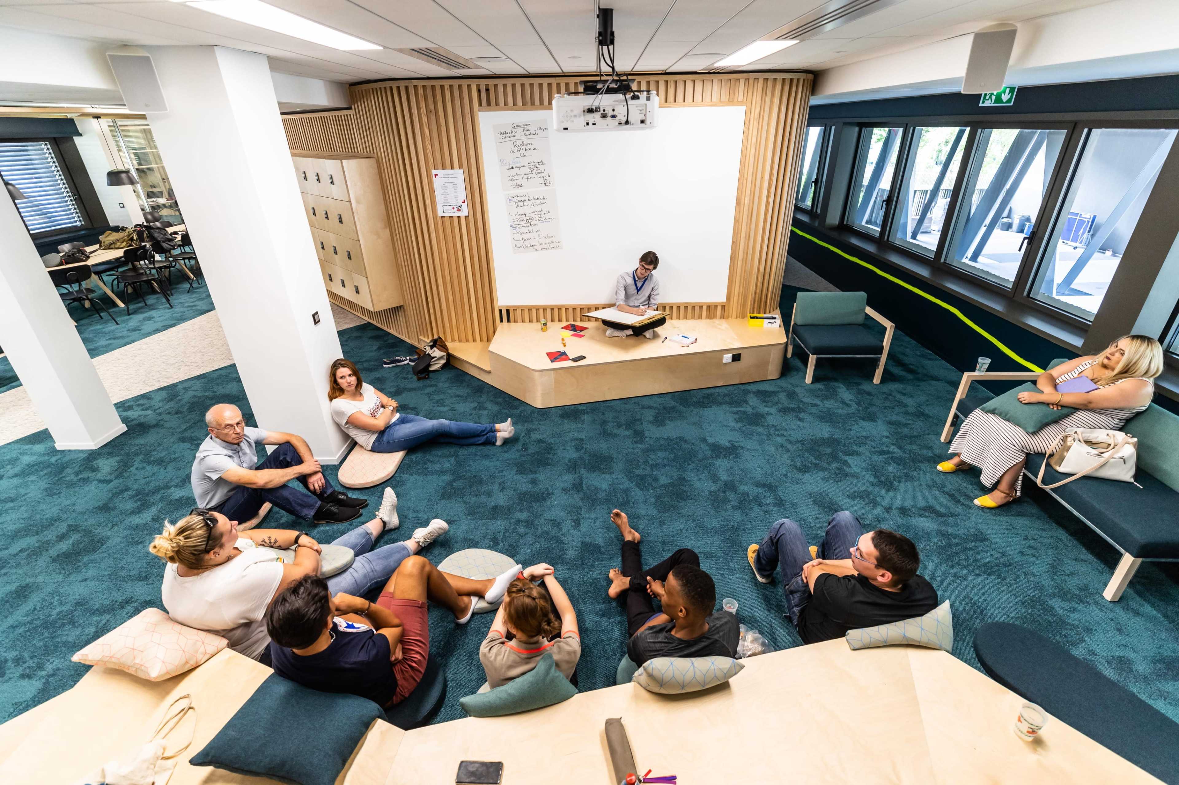 A group of people is seated on the floor and couches in a modern learning space, while a person writes on a whiteboard at the front.