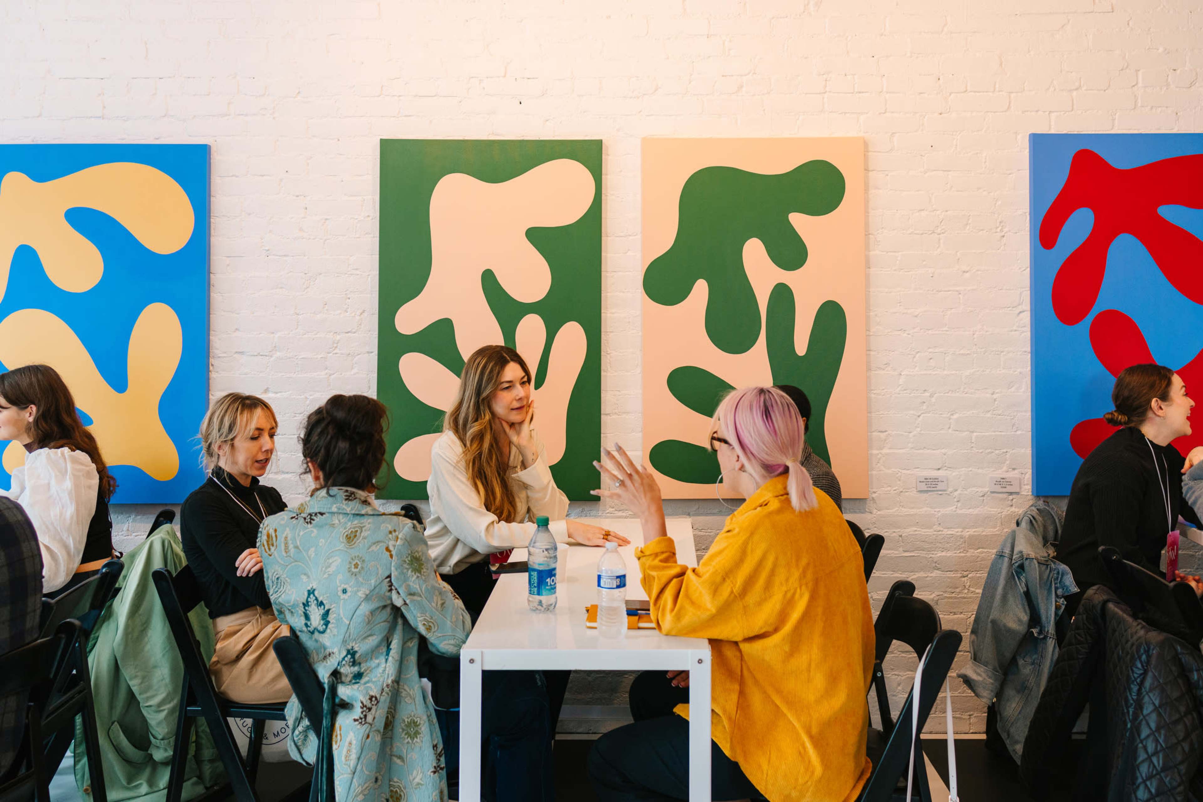 A group of people are seated at tables in a brightly lit room, with large abstract paintings on the walls behind them.