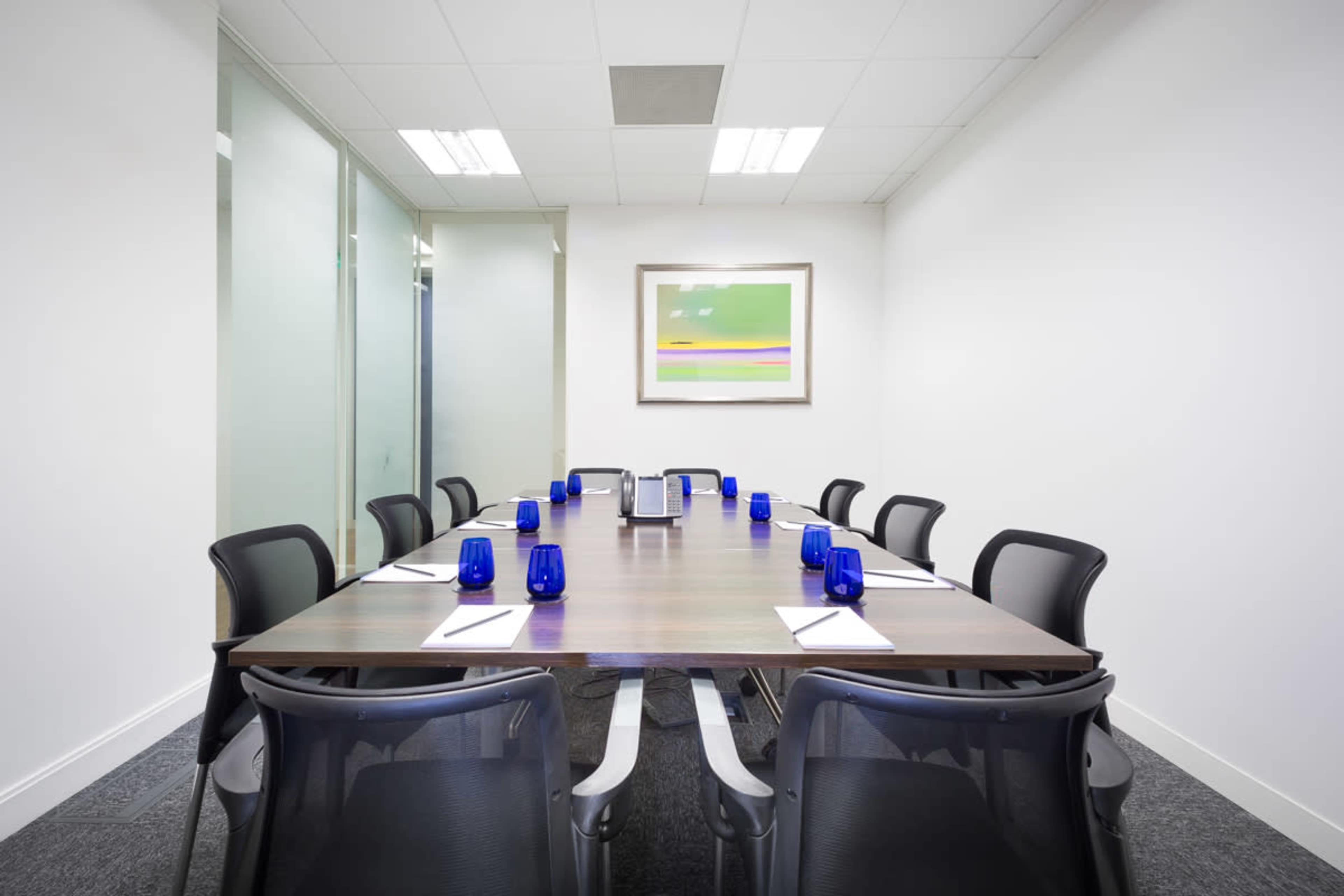 A long wooden conference table is set with blue glasses and notepads, surrounded by black chairs in a brightly lit meeting room.