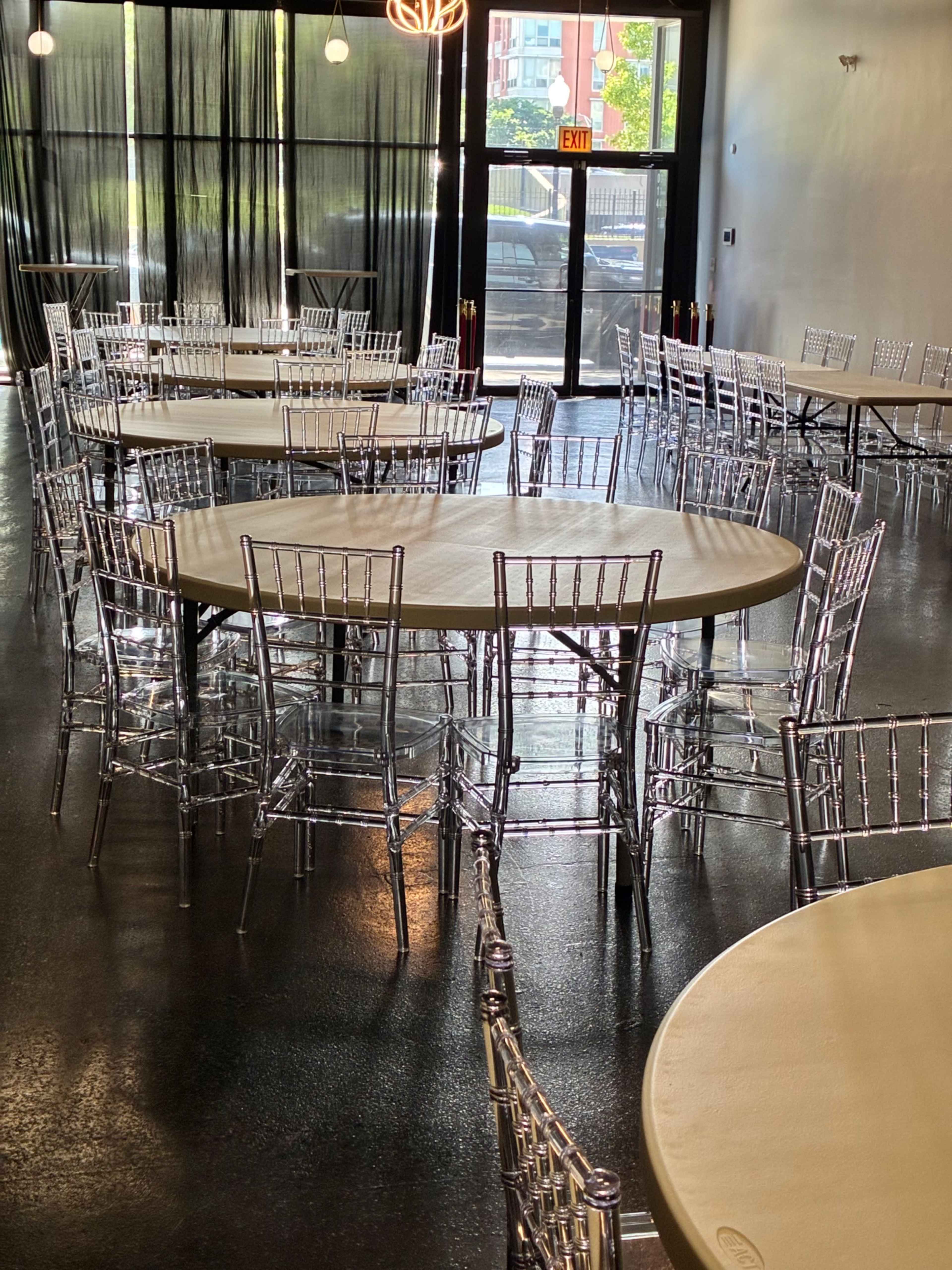 A spacious event room features several round tables surrounded by clear, chiavari-style chairs, with large windows allowing natural light to enter.
