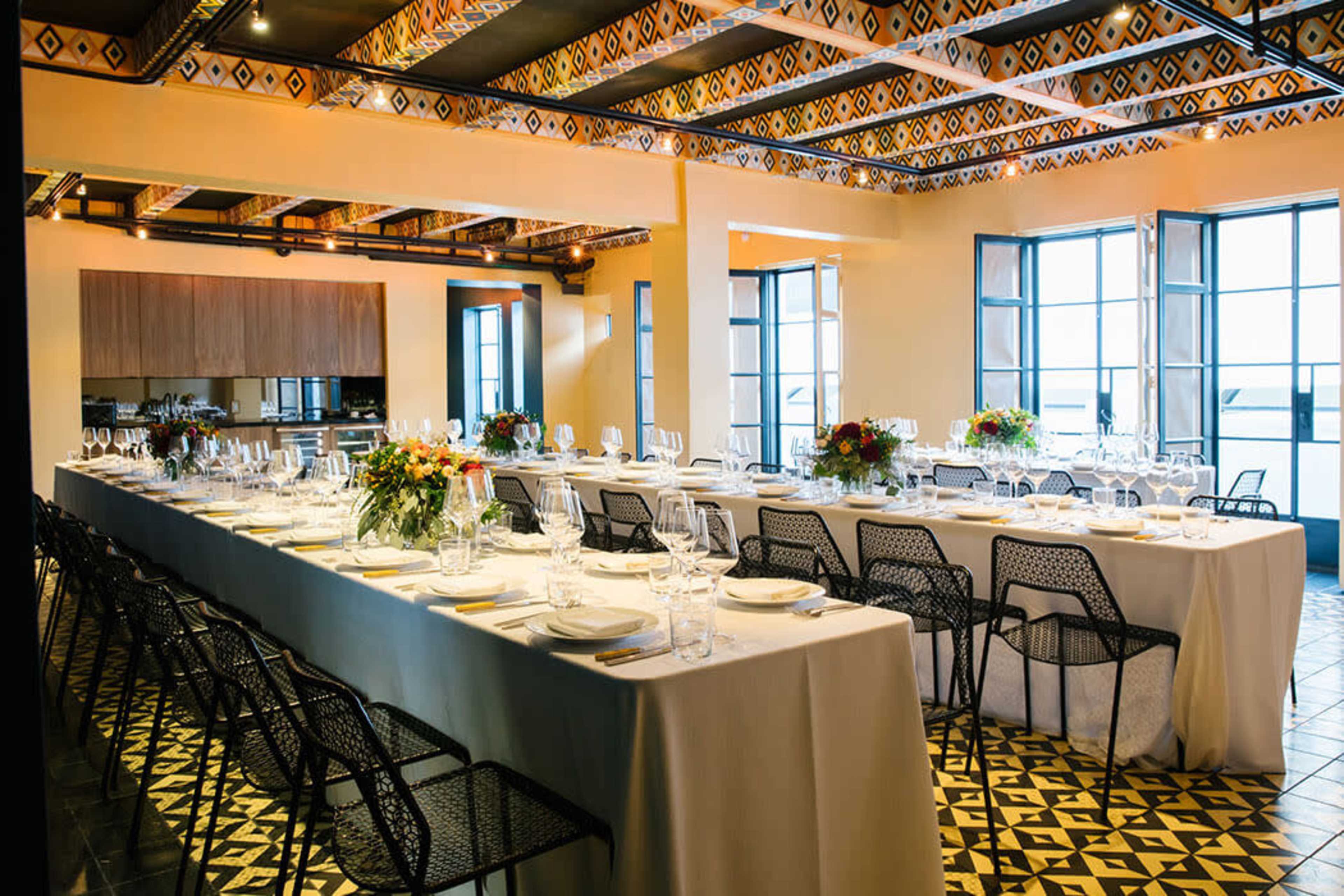 A large dining room features a long table set with tableware and floral centerpieces, surrounded by black metal chairs, under a patterned ceiling and bright windows.