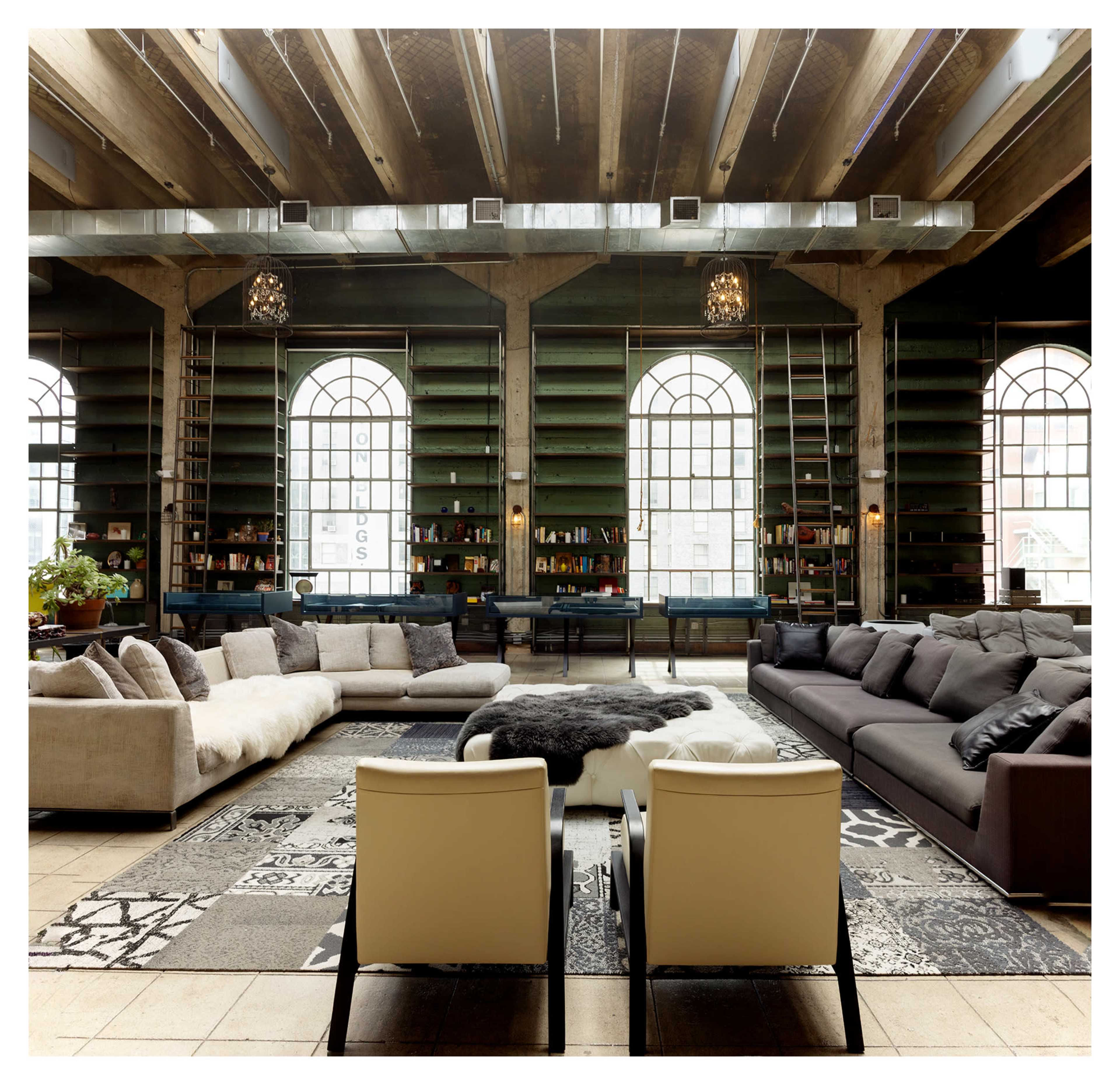 The image shows a spacious, modern interior with two large sofas facing each other, a central circular rug, and tall windows that allow natural light to fill the room.