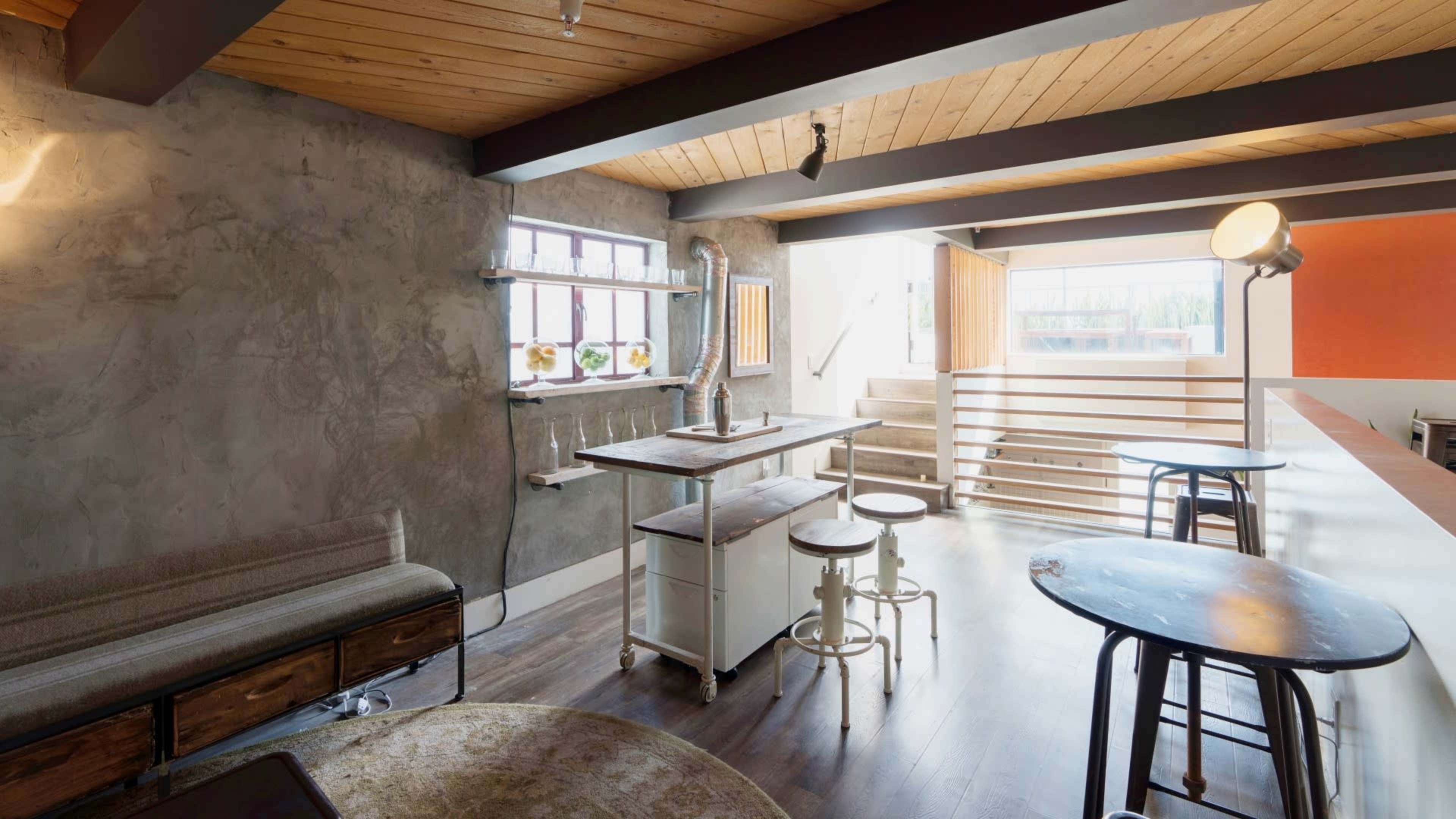 The image shows a modern interior space featuring wooden beams, a concrete wall, various tables, and natural light streaming through large windows.