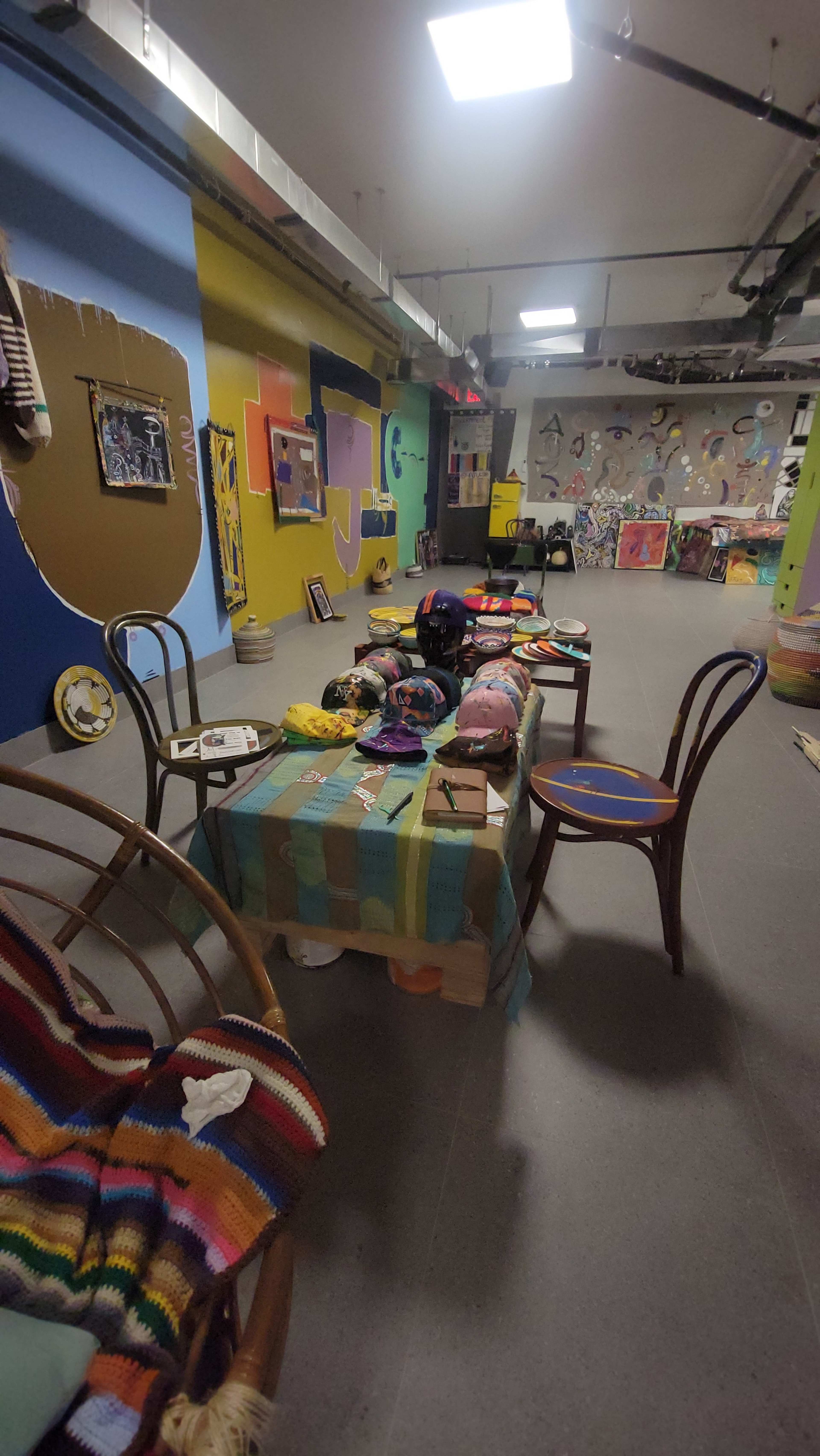 The image shows an art studio space with colorful walls, various artworks displayed, and a table set with art supplies and casual seating arrangements.