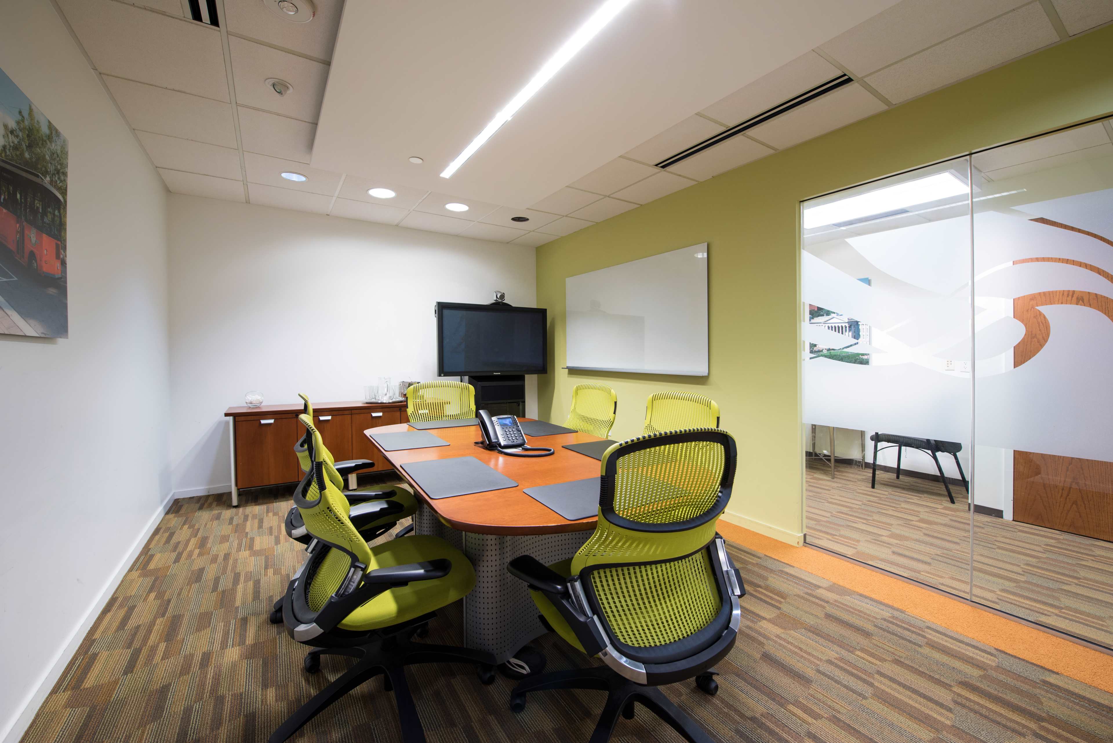 A modern conference room features a round table with six green chairs, a large screen, and a whiteboard on the wall.