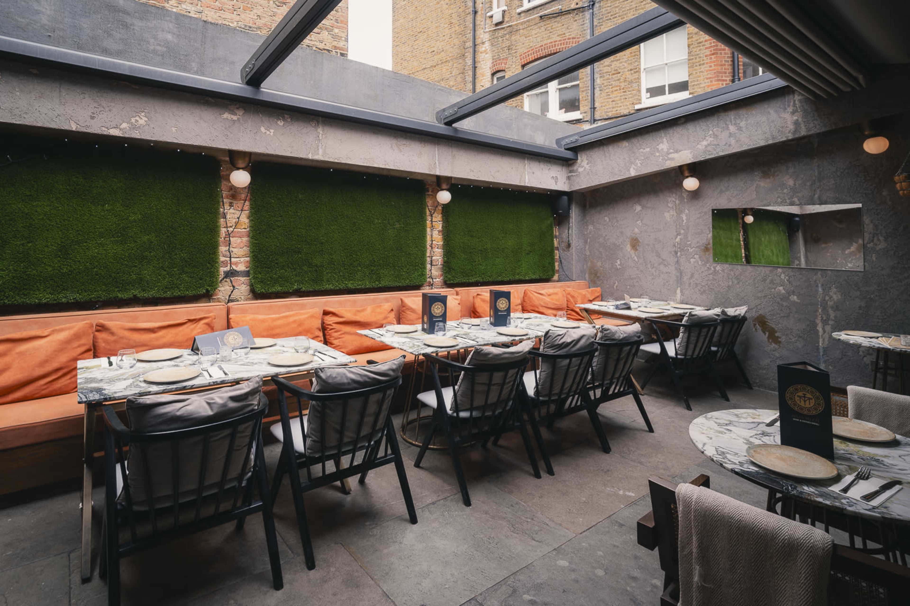 The image shows a modern restaurant with a seating area featuring marble tables, black chairs, and green moss-covered walls under a glass roof.