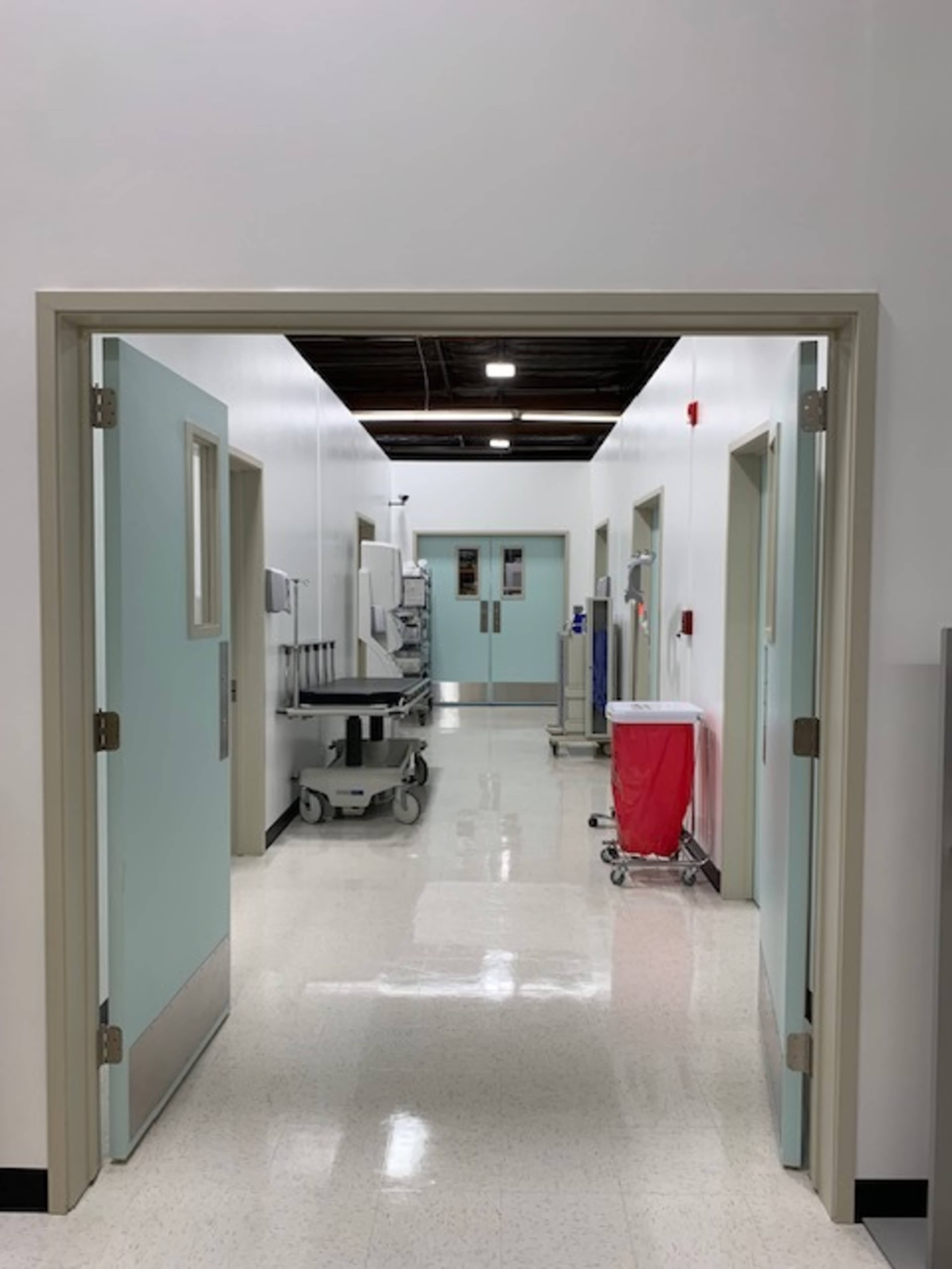 A clean, well-lit corridor in a medical facility, with several doors on either side and equipment visible in the background.