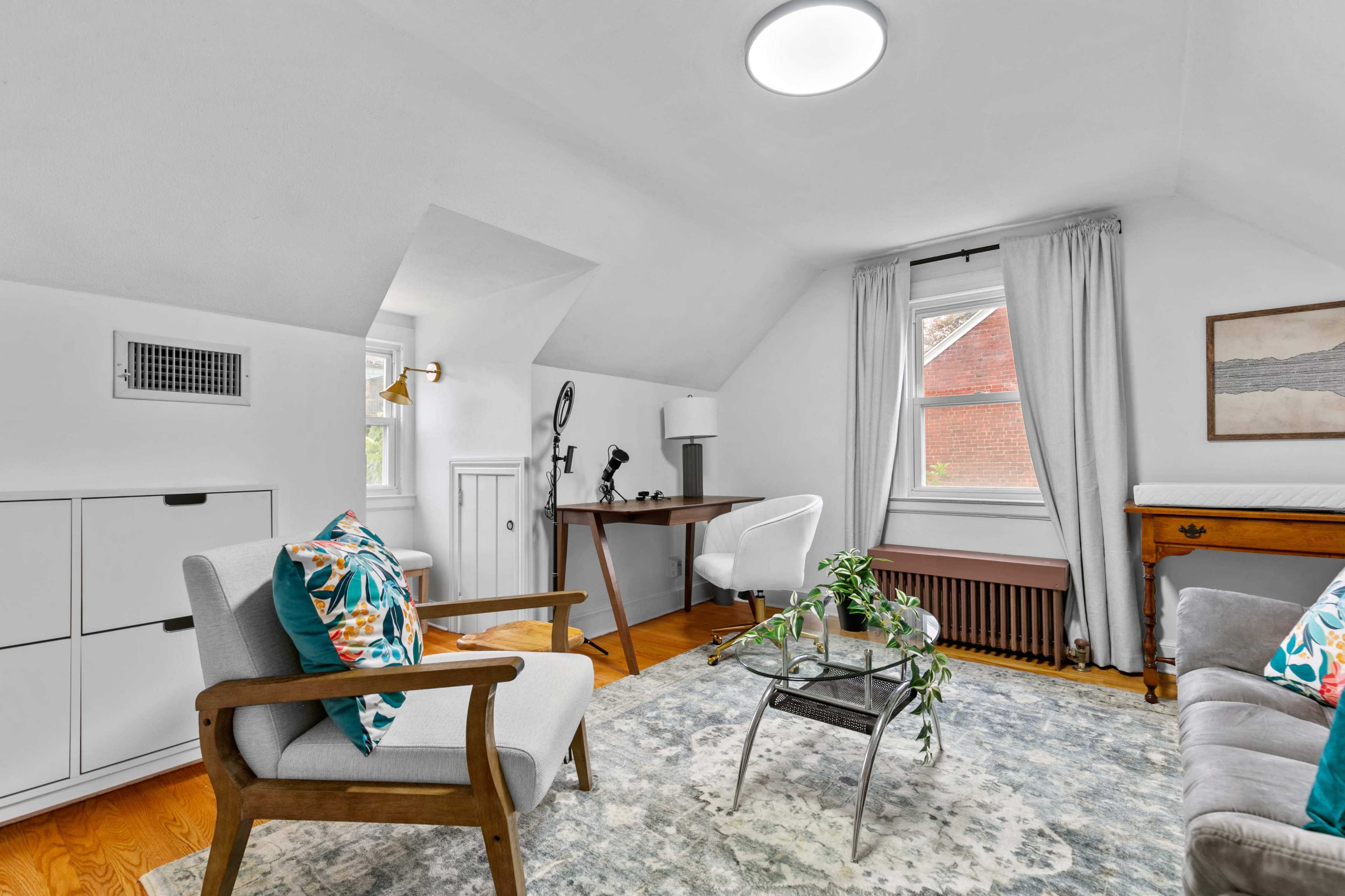 A bright, modern sitting room features a mix of furniture including a gray armchair, a desk, and a comfortable sofa, all arranged around a decorative rug and a small plant.