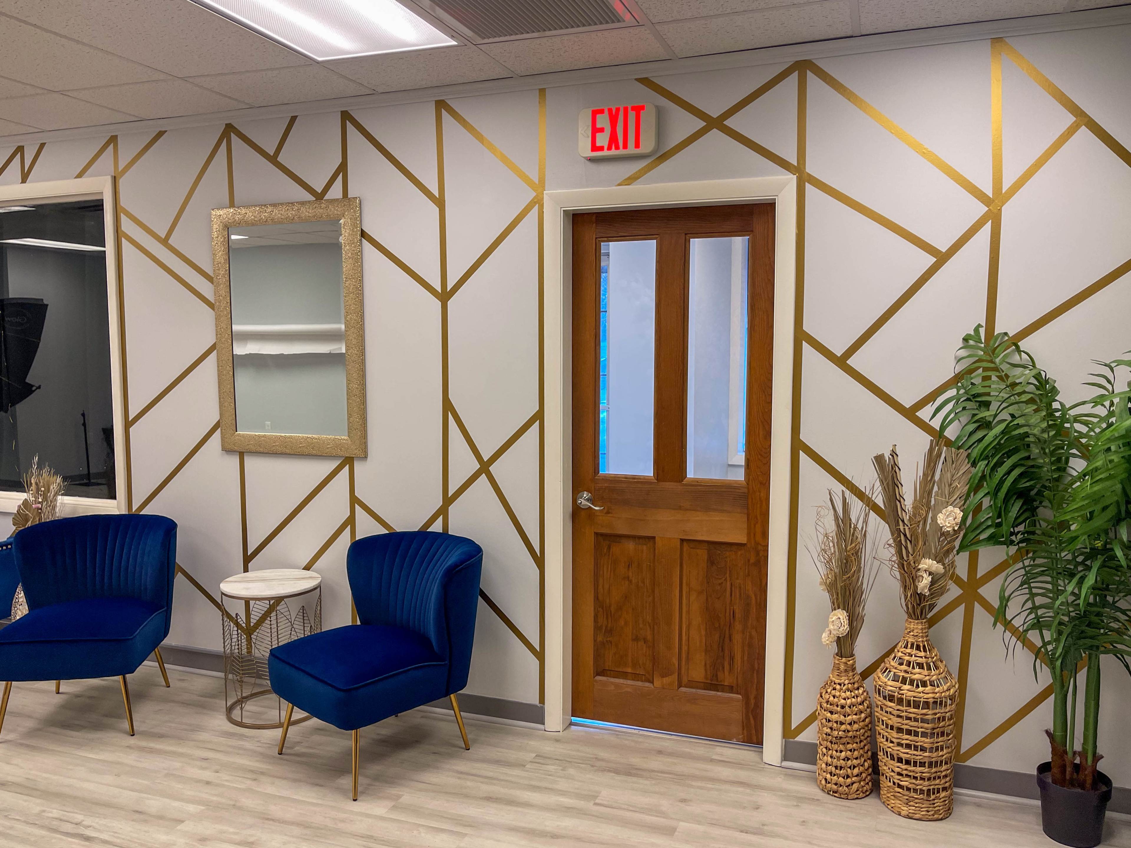 A modern waiting area with blue upholstered chairs, a decorative mirror, and a door framed by geometric gold lines on the wall.