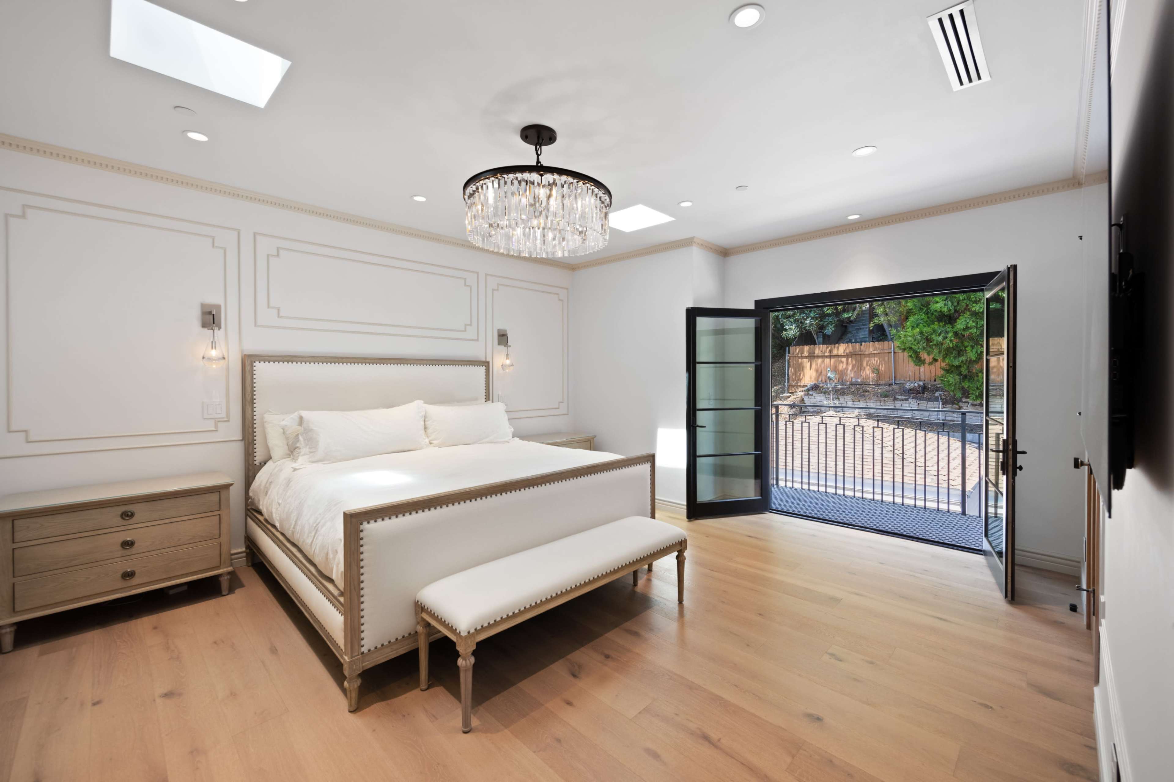 The bedroom features a bed with a bench at its foot, a chandelier overhead, and a large glass door that opens to an outdoor area.