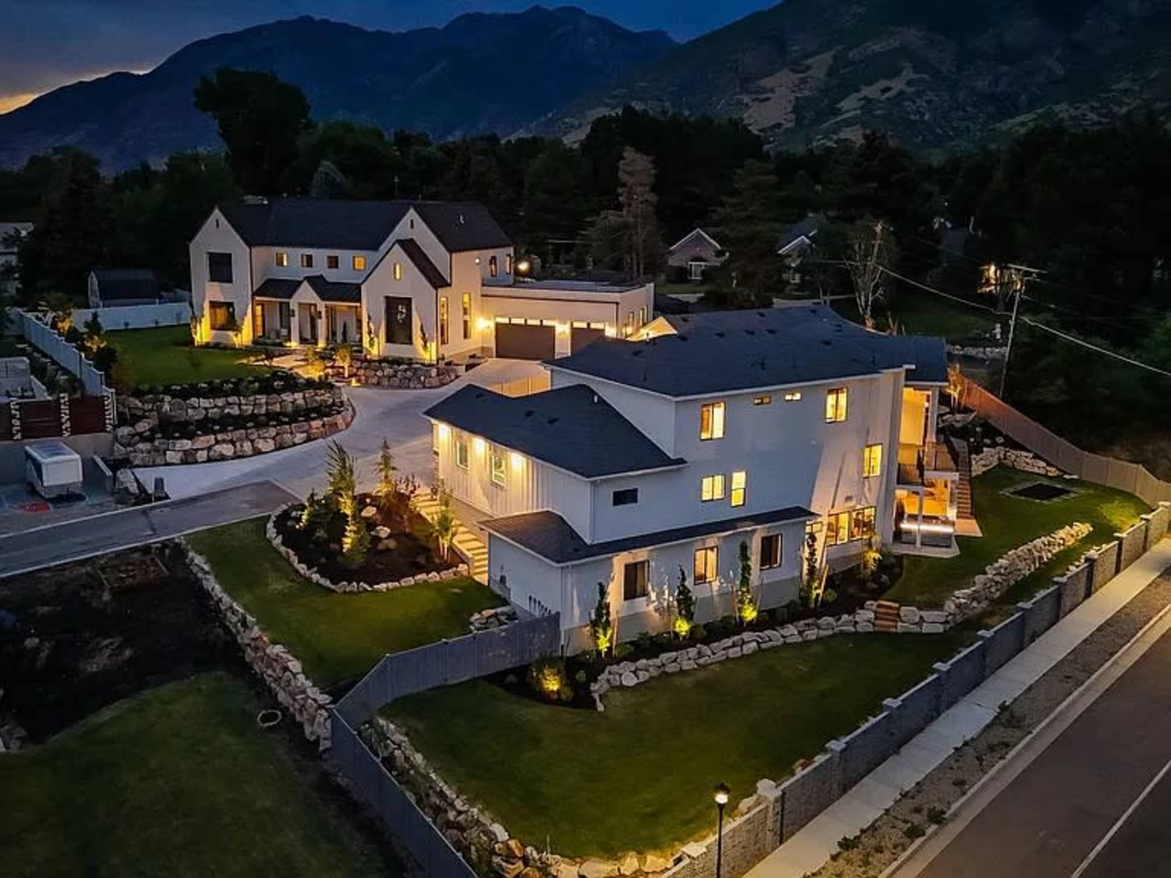 The image shows a modern residential complex with multiple buildings, landscaped gardens, and illuminated exteriors nestled against a mountainous backdrop during twilight.